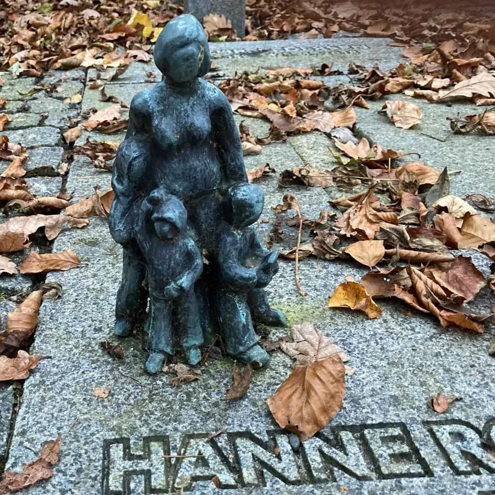 A bronze statue of a woman with a small child, placed on a stone surface with fallen autumn leaves around. The statue has the word 'HANNER' engraved at the base.