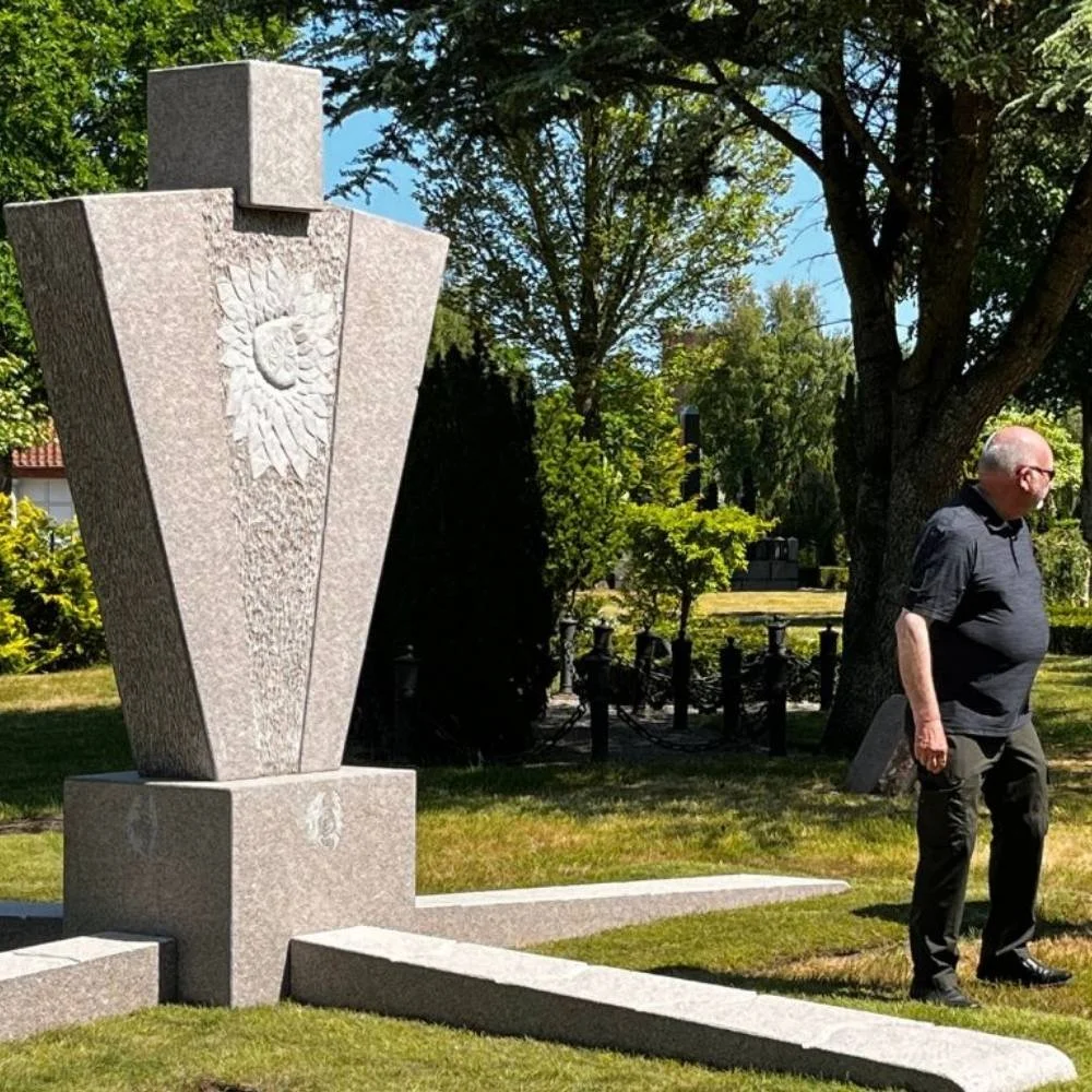 A granite monument with a carved sun design in a park, and a man dressed in black walking nearby under trees on a sunny day.