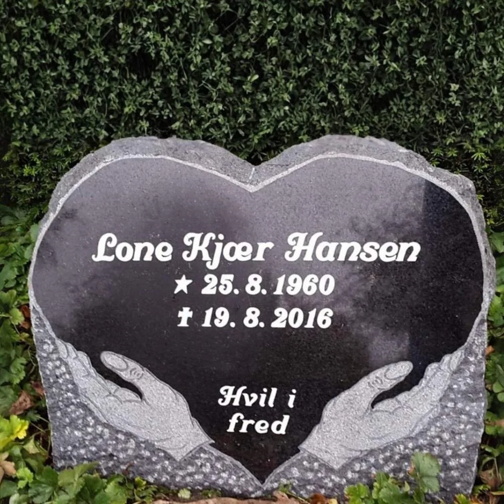 A black tombstone with a carved heart shape and two hands at the bottom, surrounded by greenery. The tombstone bears the inscription 'Lone Kjaer Hansen, born August 25, 1960, died August 19, 2016, with the Danish phrase 'Hvil i fred' (rest in peace).