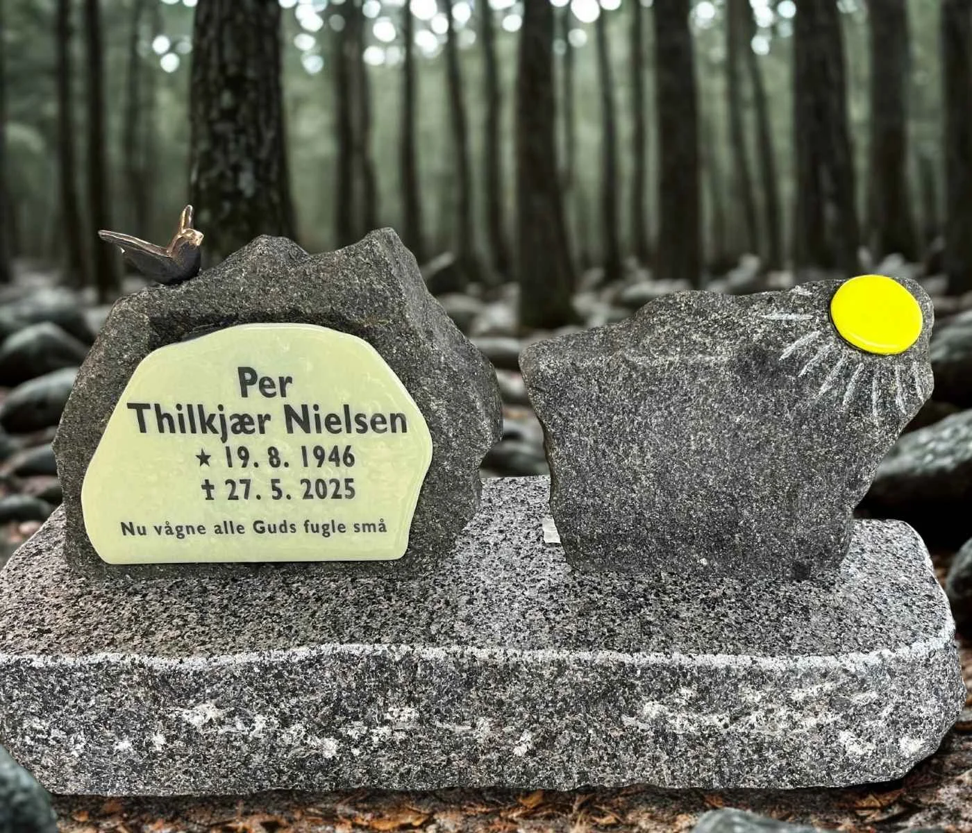 A memorial stone with a small plaque for Thillkjær Nielsen, displaying birth and death dates, with a small bird figurine on top and a yellow sticker on a second stone, set in a forest.