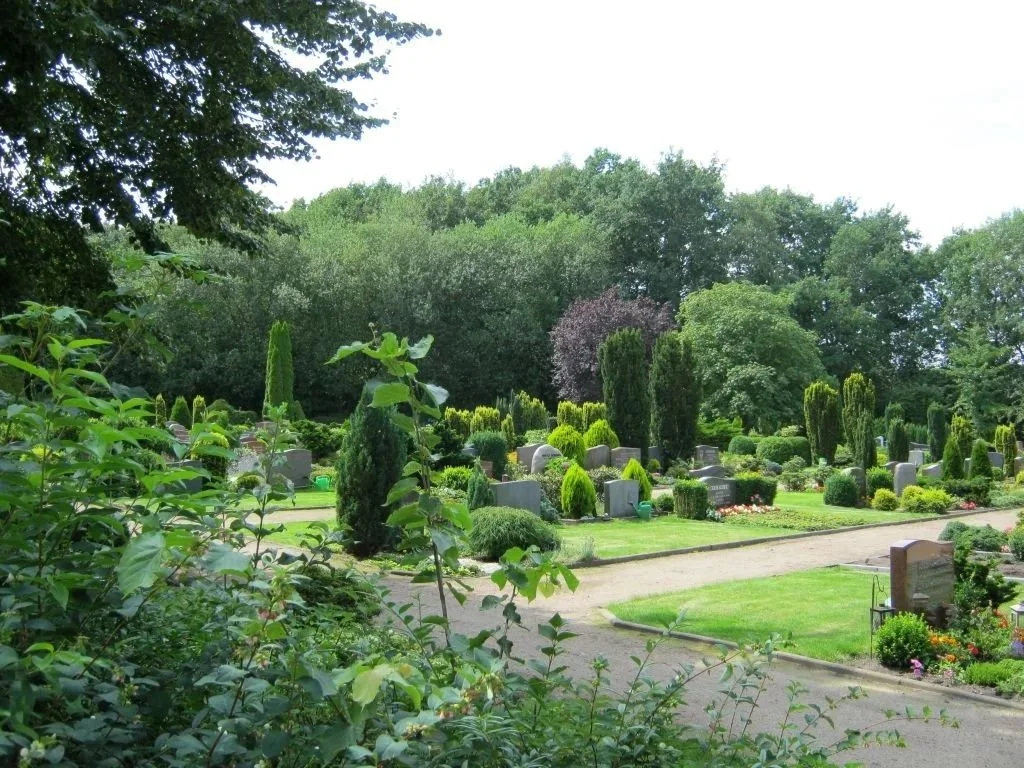 A peaceful cemetery scene with numerous headstones surrounded by lush green bushes and tall trees.