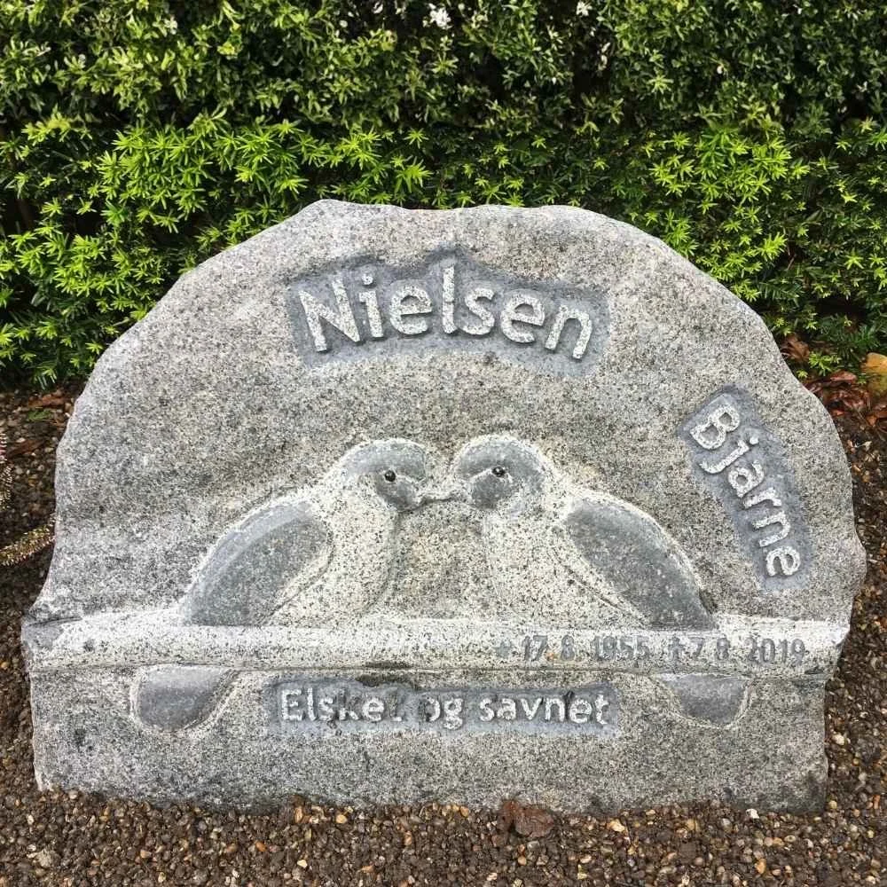 Gravestone with an engraved image of two birds facing each other, surrounded by green shrubbery. The inscription reads "Nielsen Bjørne" at the top, with dates "17.6.1953 - 27.8.2019" below, and the words "Elsker og savner" at the bottom, which means 