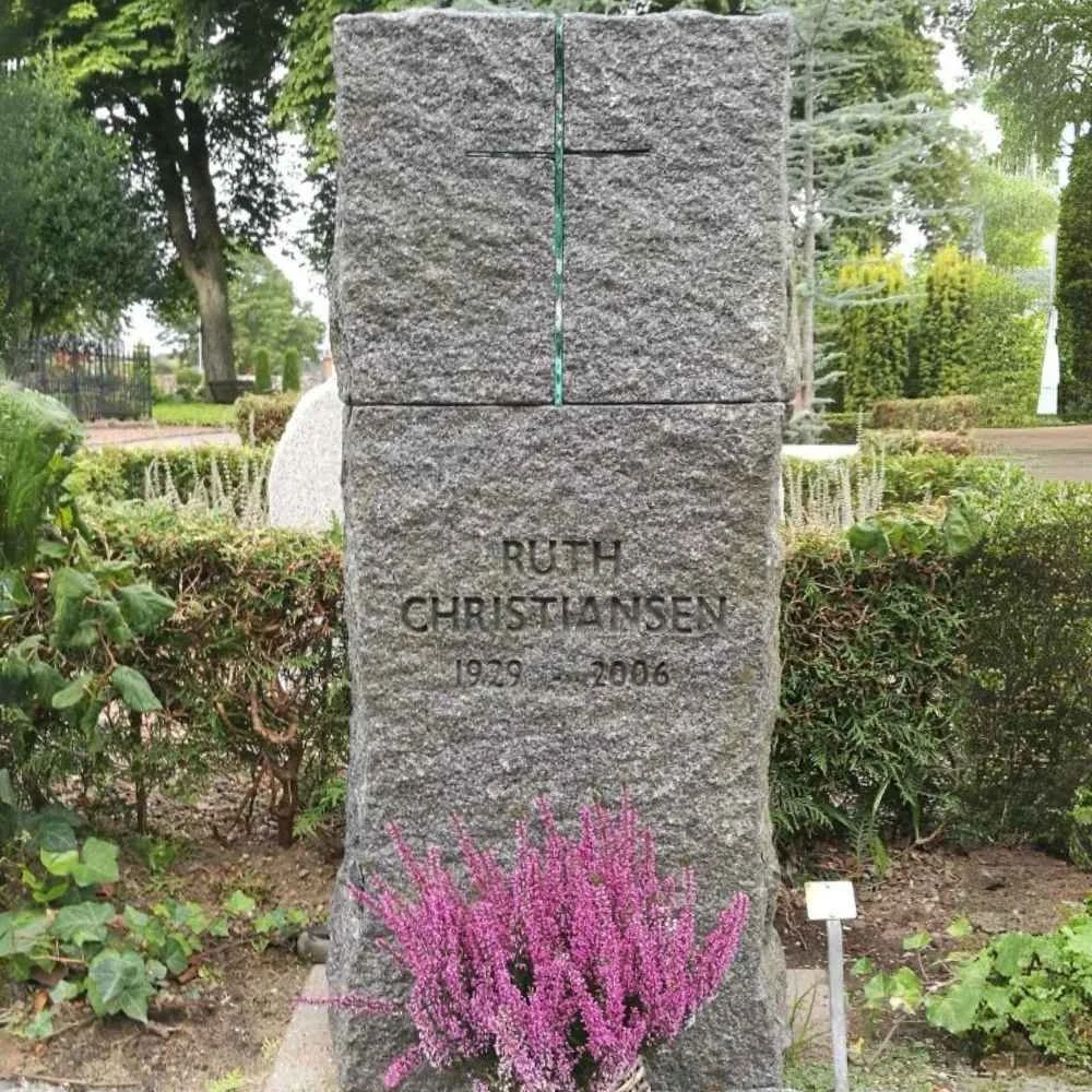 Granite memorial stone engraved with the name Ruth Christiansen, with birth year 1929 and death year 2006, decorated with pink flowers at the base, situated outdoors in a garden with trees and shrubs.