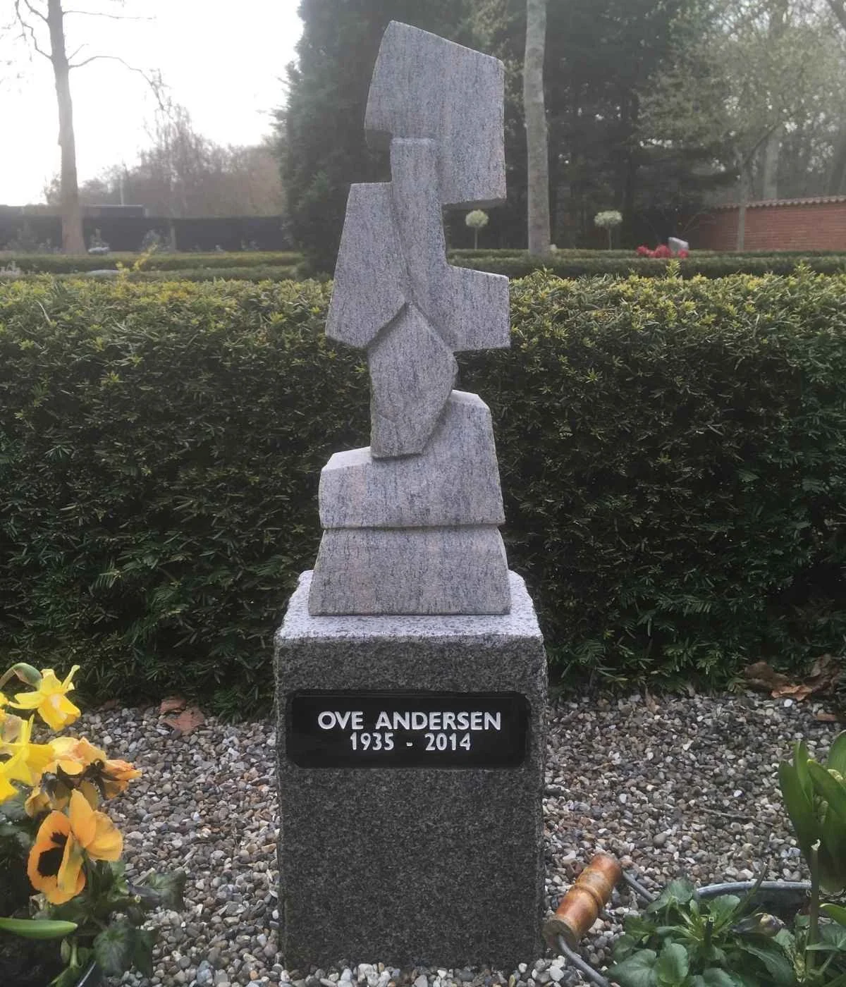 Memorial stone with abstract sculpture on top, dedicated to Ove Andersen (1935-2014), situated in a park with greenery and flowers.