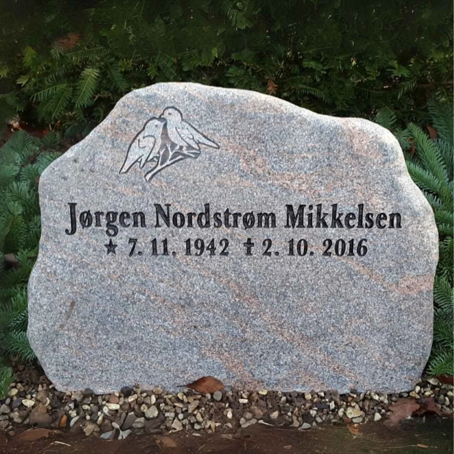 A gray granite headstone with an engraving of two birds on a branch at the top left corner. The headstone has the name 'Jorgen Nordstrom Mikkelsen' engraved in black, along with birth and death dates: November 7, 1942, and October 2, 2016. The headst