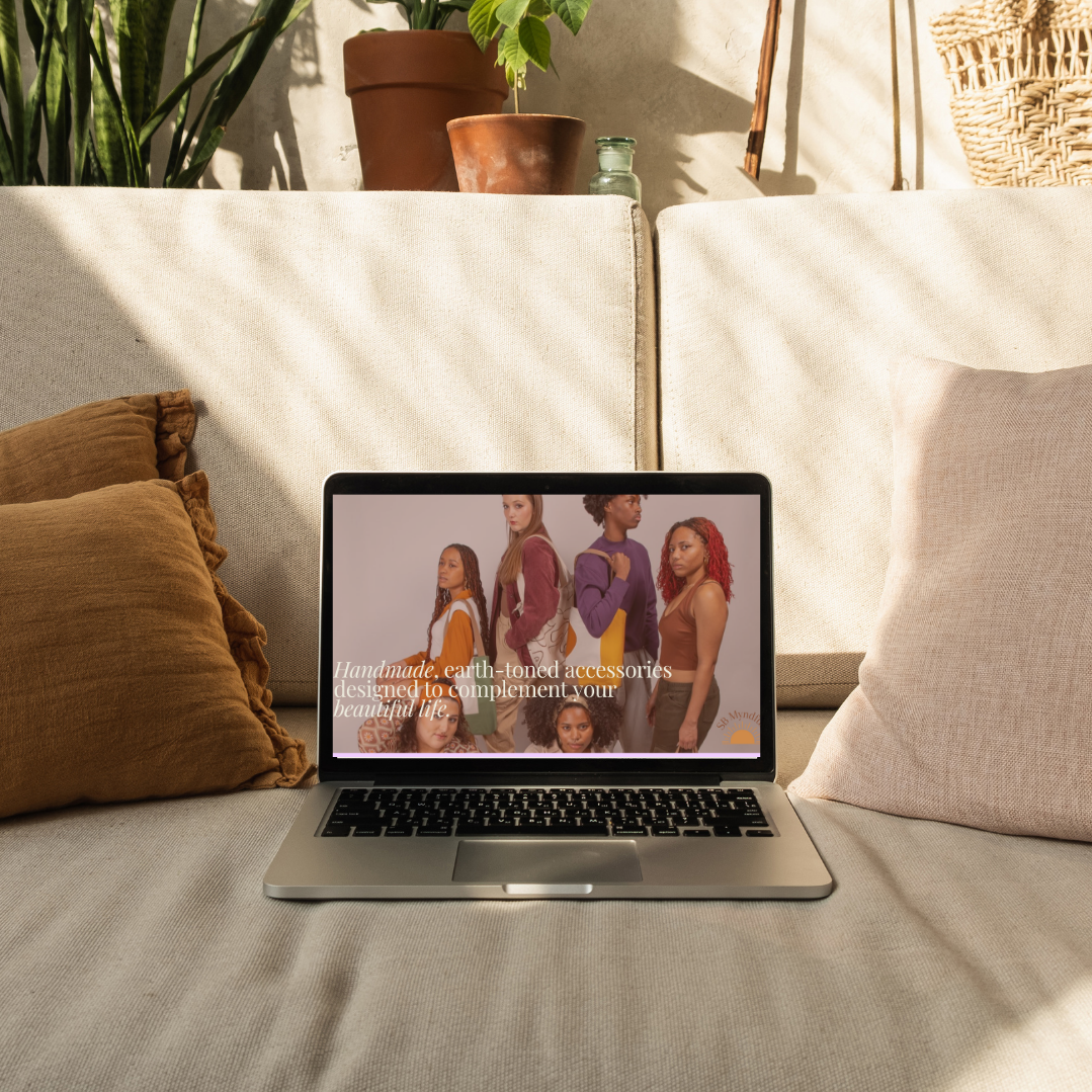Silver laptop on a beige couch displaying a fashion website with models and text about earth-toned accessories. The background includes potted plants and woven décor.
