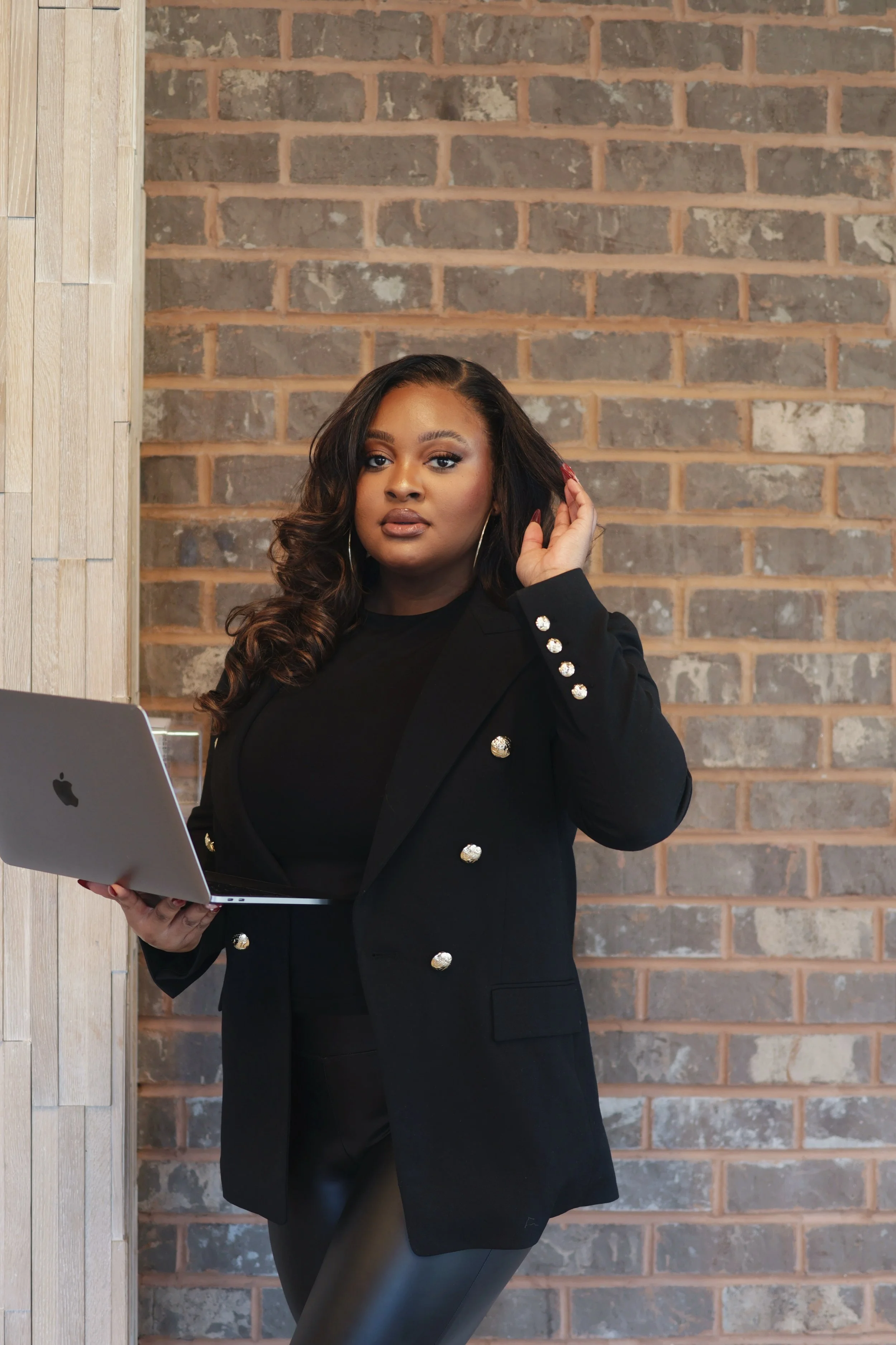 A confident woman with dark hair and makeup, wearing a black blazer with decorative buttons, holding a silver MacBook, standing against a brick wall in an indoor setting.