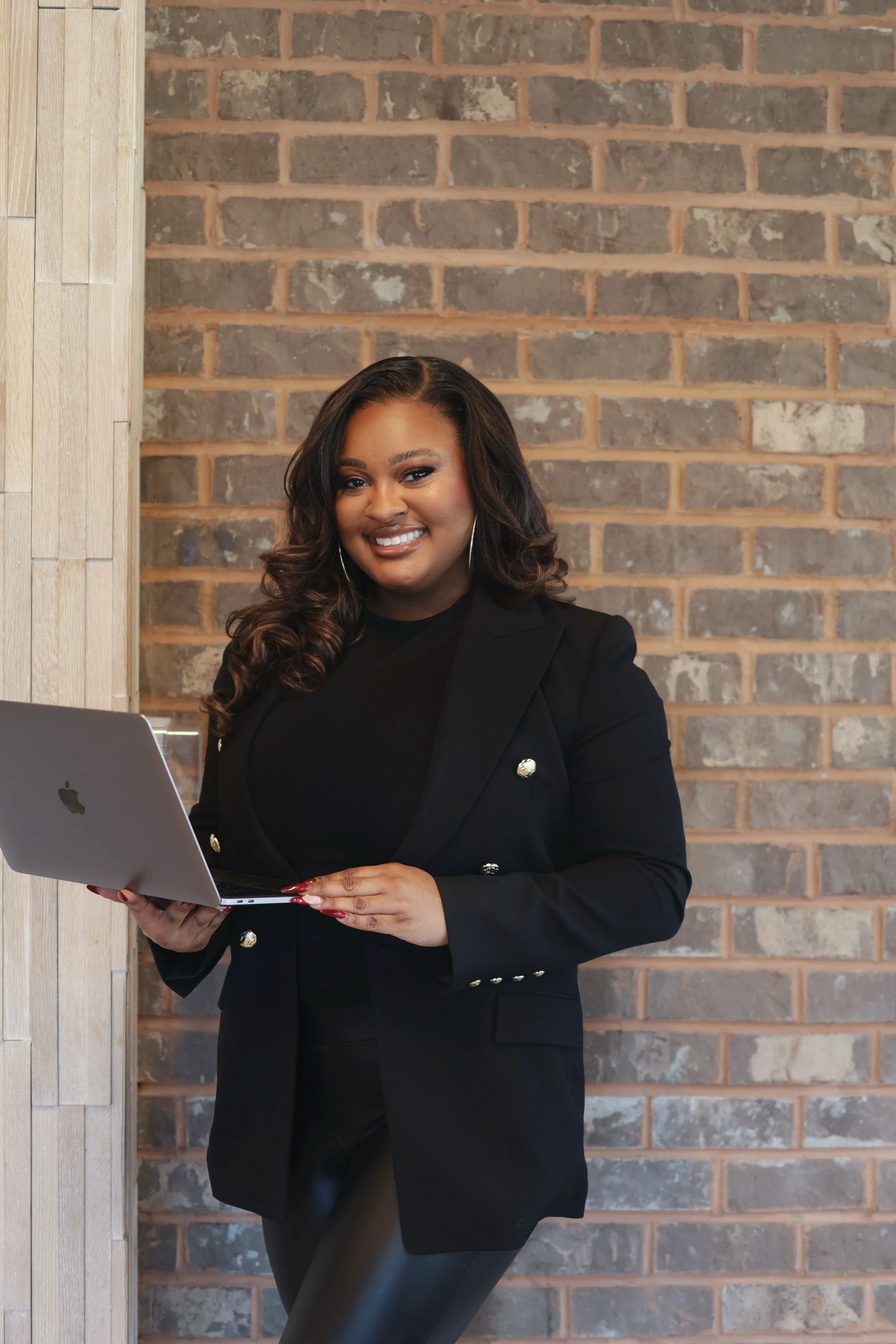 A woman standing in front of a brick wall, smiling, holding a silver MacBook laptop, dressed in a black blazer and pants.