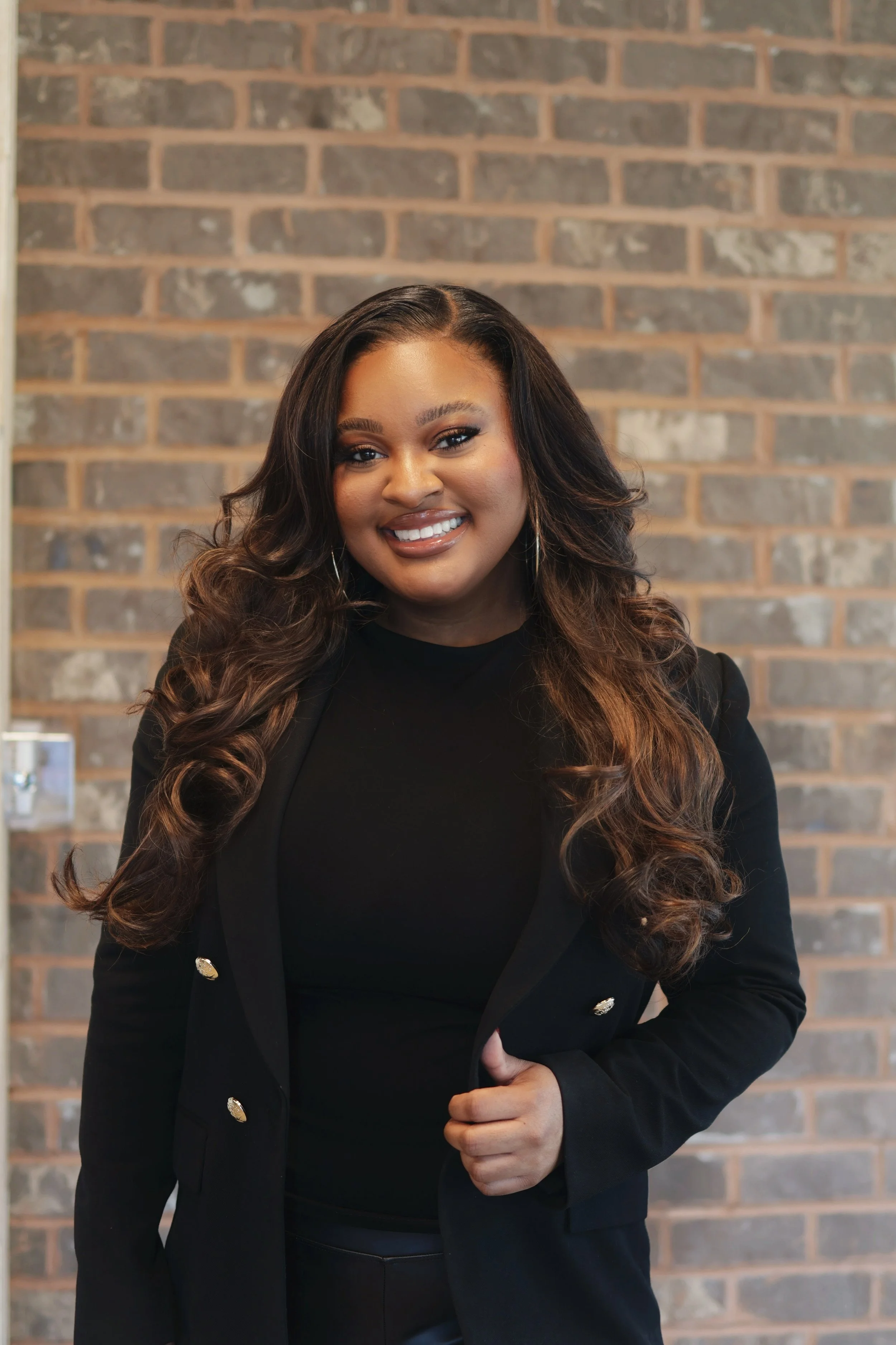 A smiling woman with long, curly hair, wearing a black blazer over a black top, standing in front of a brick wall.