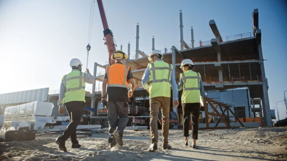 Construction workers in safety vests and helmets inspecting a building site with cranes and unfinished structure.