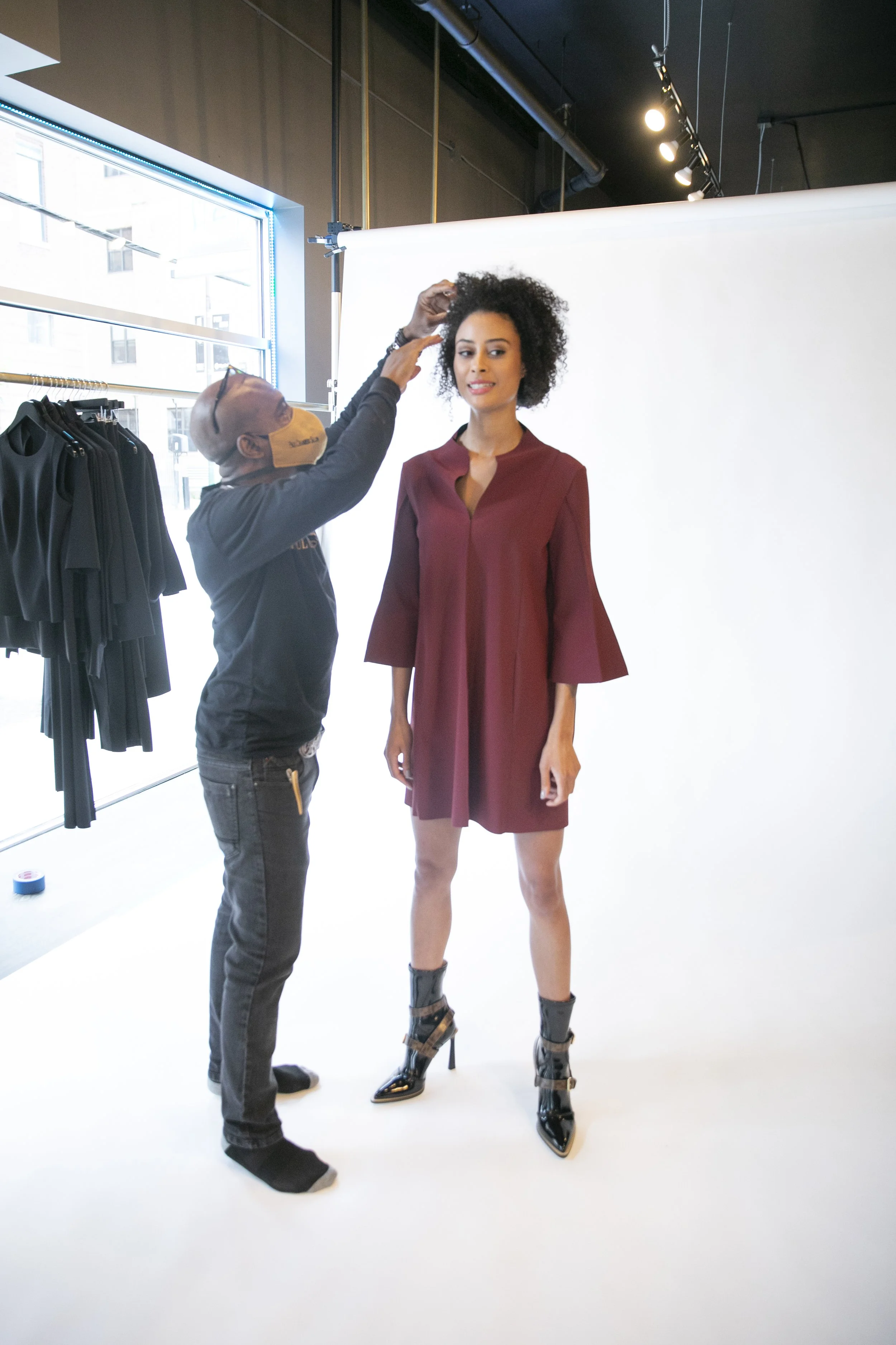 A fashion photoshoot studio with a model wearing a maroon dress and black high-heeled boots, while a photographer adjusts the model's hair. The studio has a white backdrop, rack of black clothing, and large window letting in natural light.