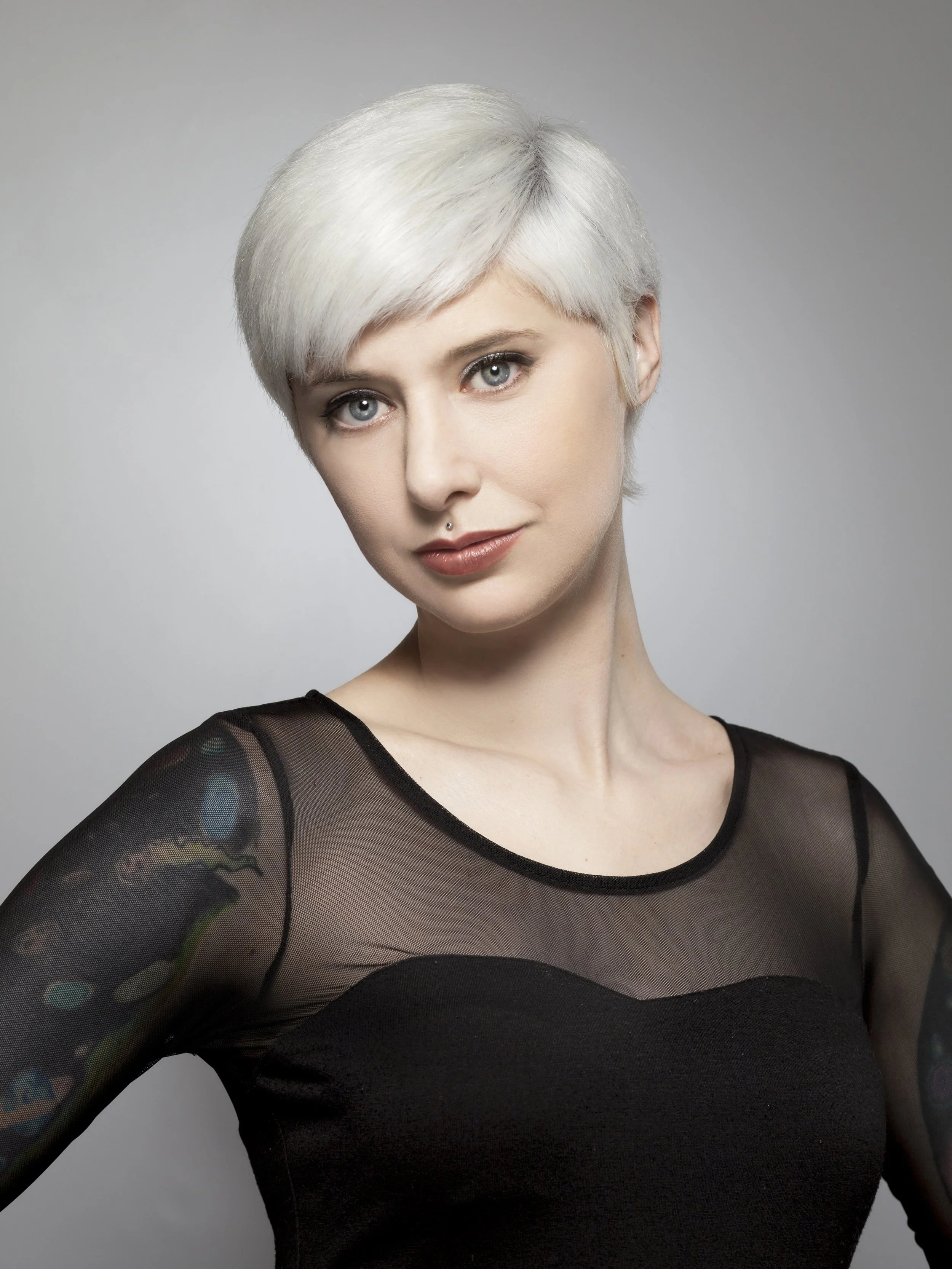 Portrait of a woman with short, platinum blonde hair, wearing a black sheer top with embroidered sleeves, looking directly at the camera with a slight smile.