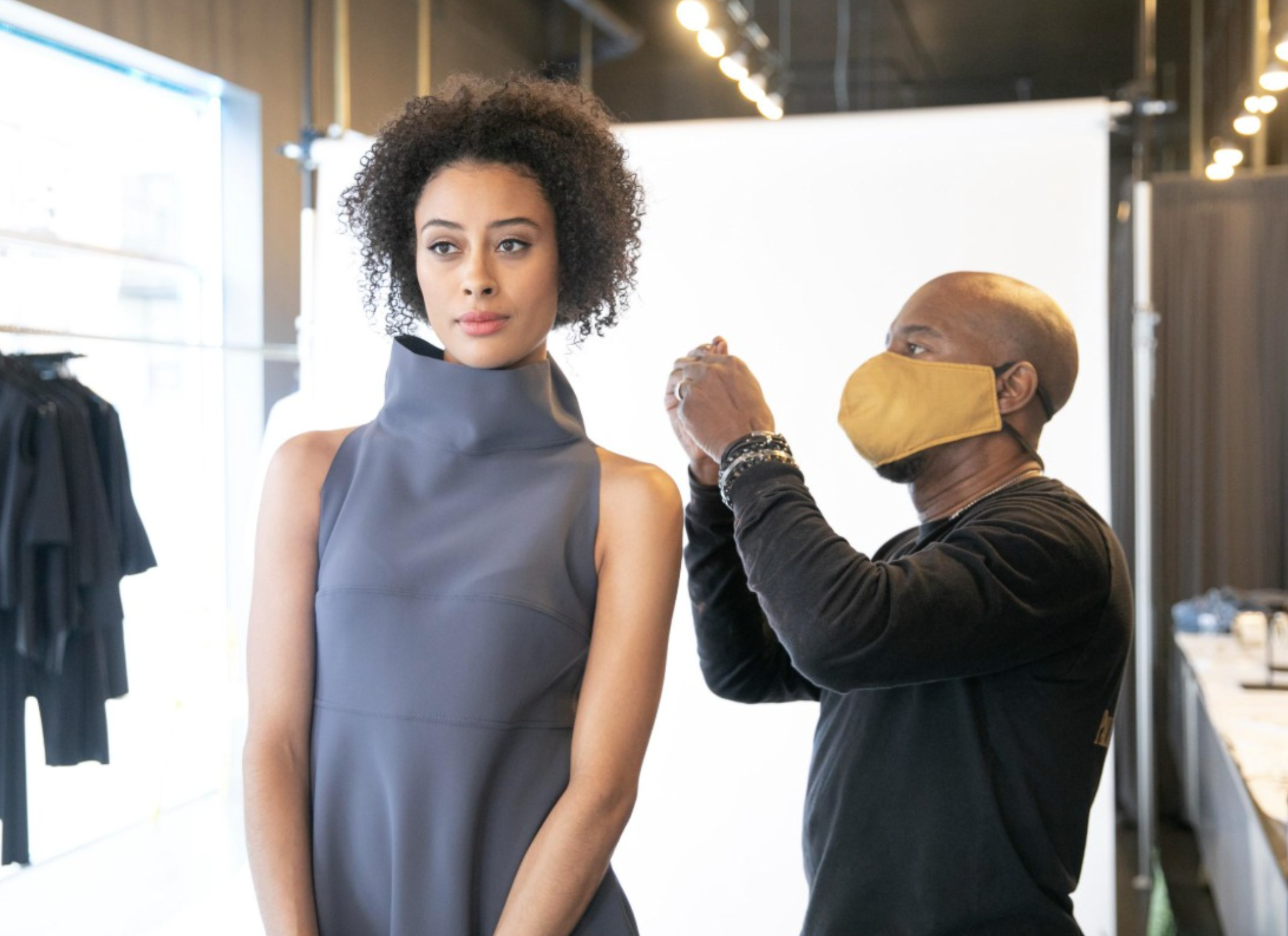 A woman with curly hair models a sleeveless, high-collared gray dress while a stylist adjusts her outfit, both in a well-lit fashion studio.