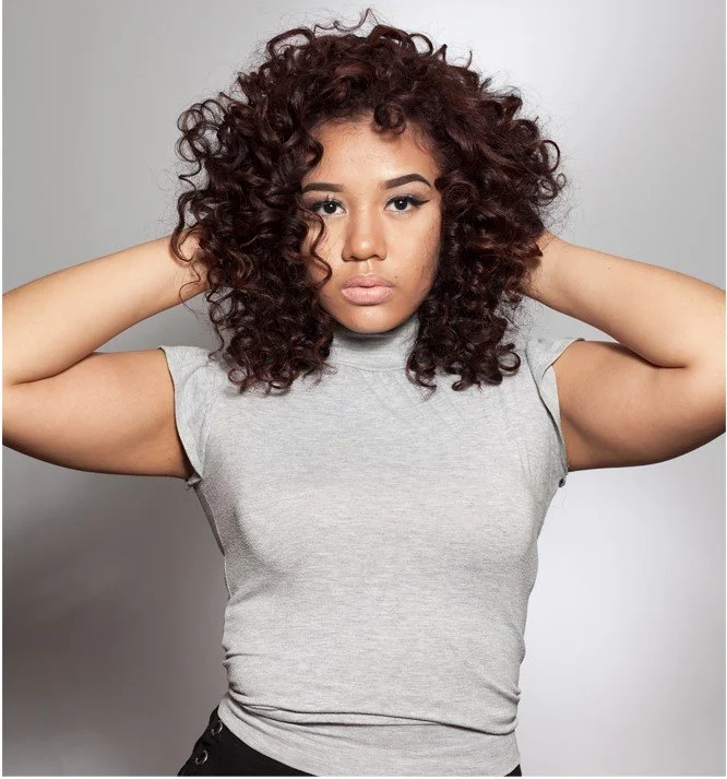 A young woman with curly hair wearing a gray t-shirt, holding her hands behind her head, looking directly at the camera, against a plain gray background.