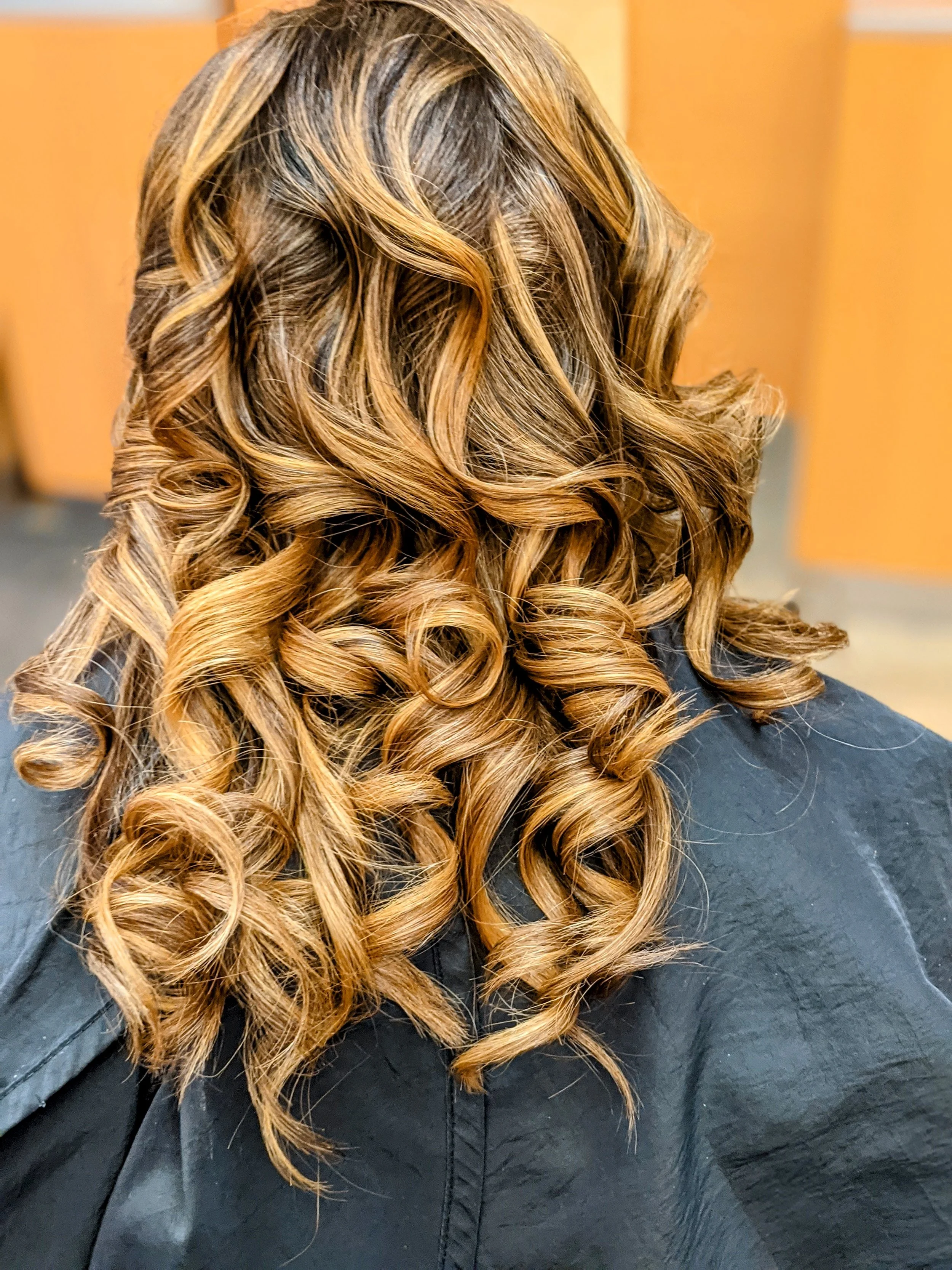 Back view of a woman with long, styled, curly hair in shades of blonde and light brown, wearing a black cape, in a salon setting.