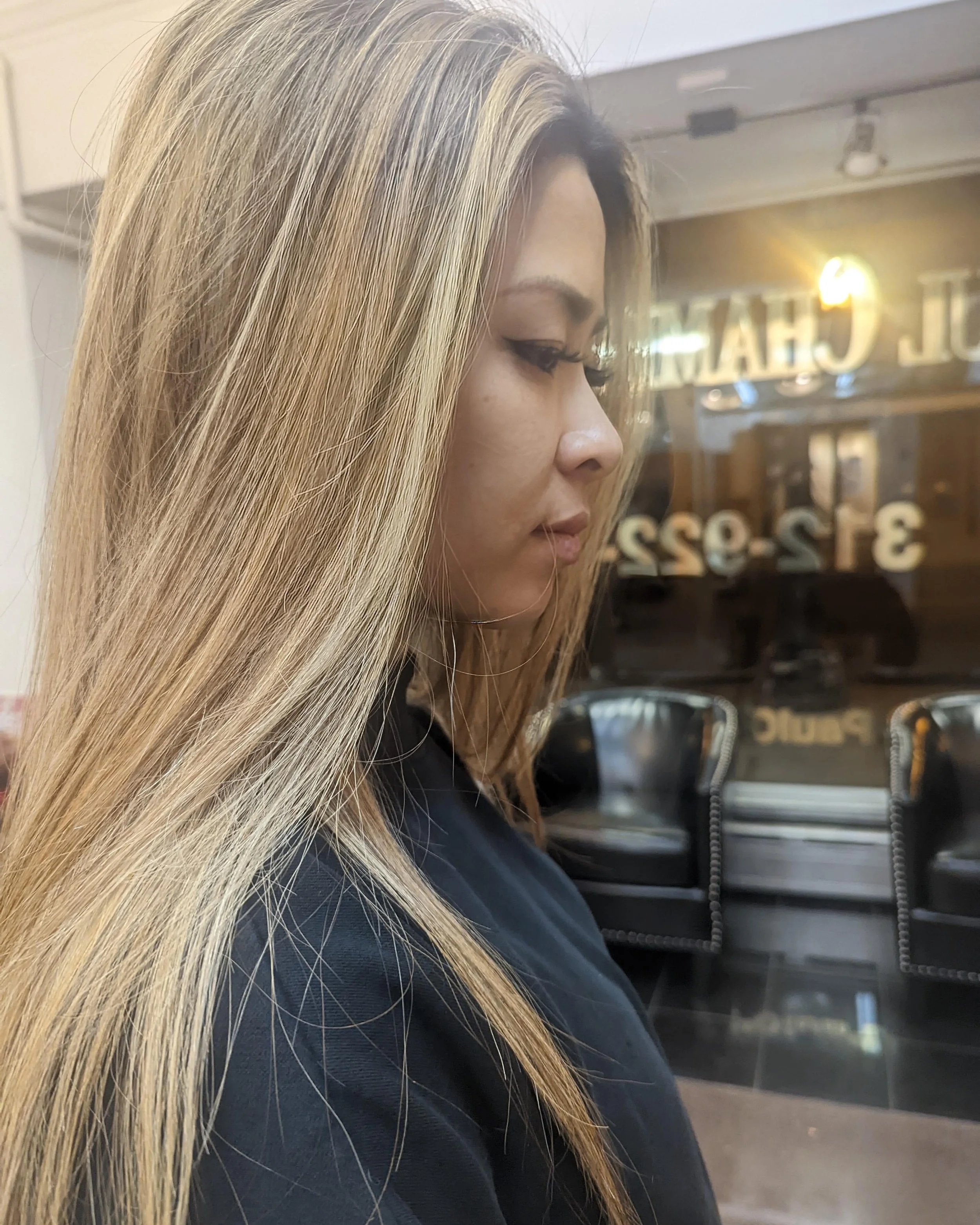 A woman with long blonde hair sitting indoors, looking downwards with a serious expression, in front of a glass window with reflections of street lights and a sign.