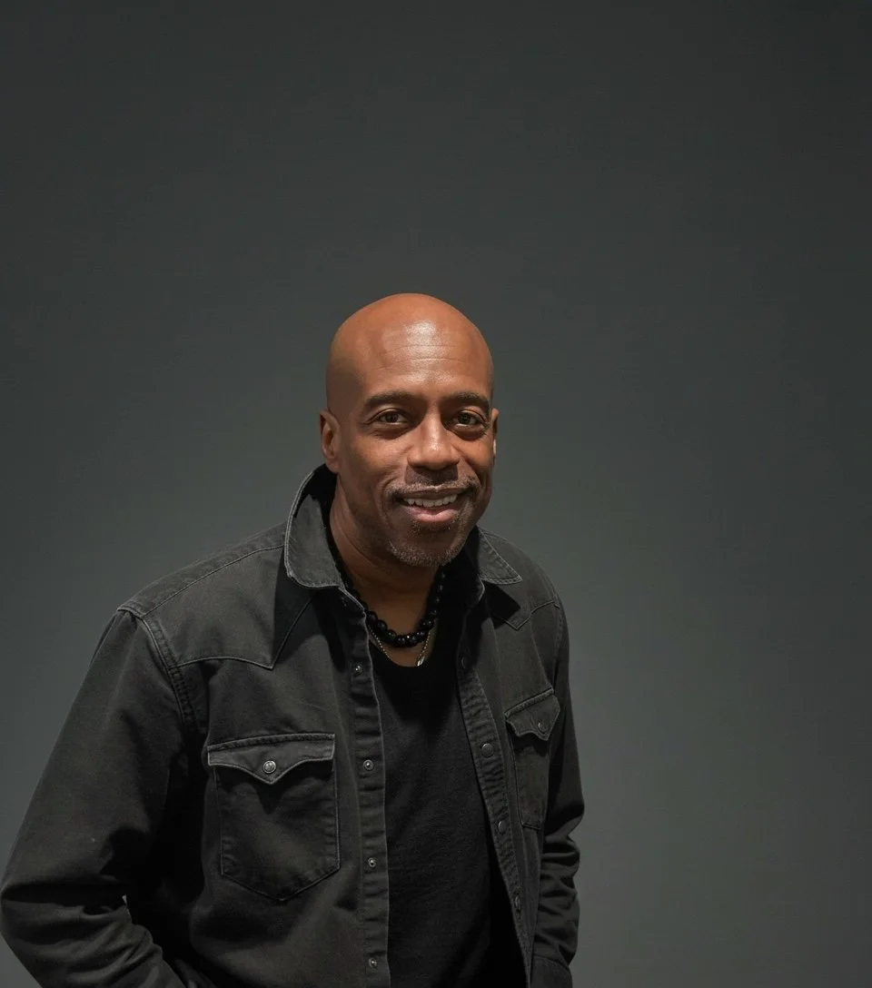 Man with dark skin, bald head, black shirt, black beaded necklace, black jacket, smiling against a plain dark background.