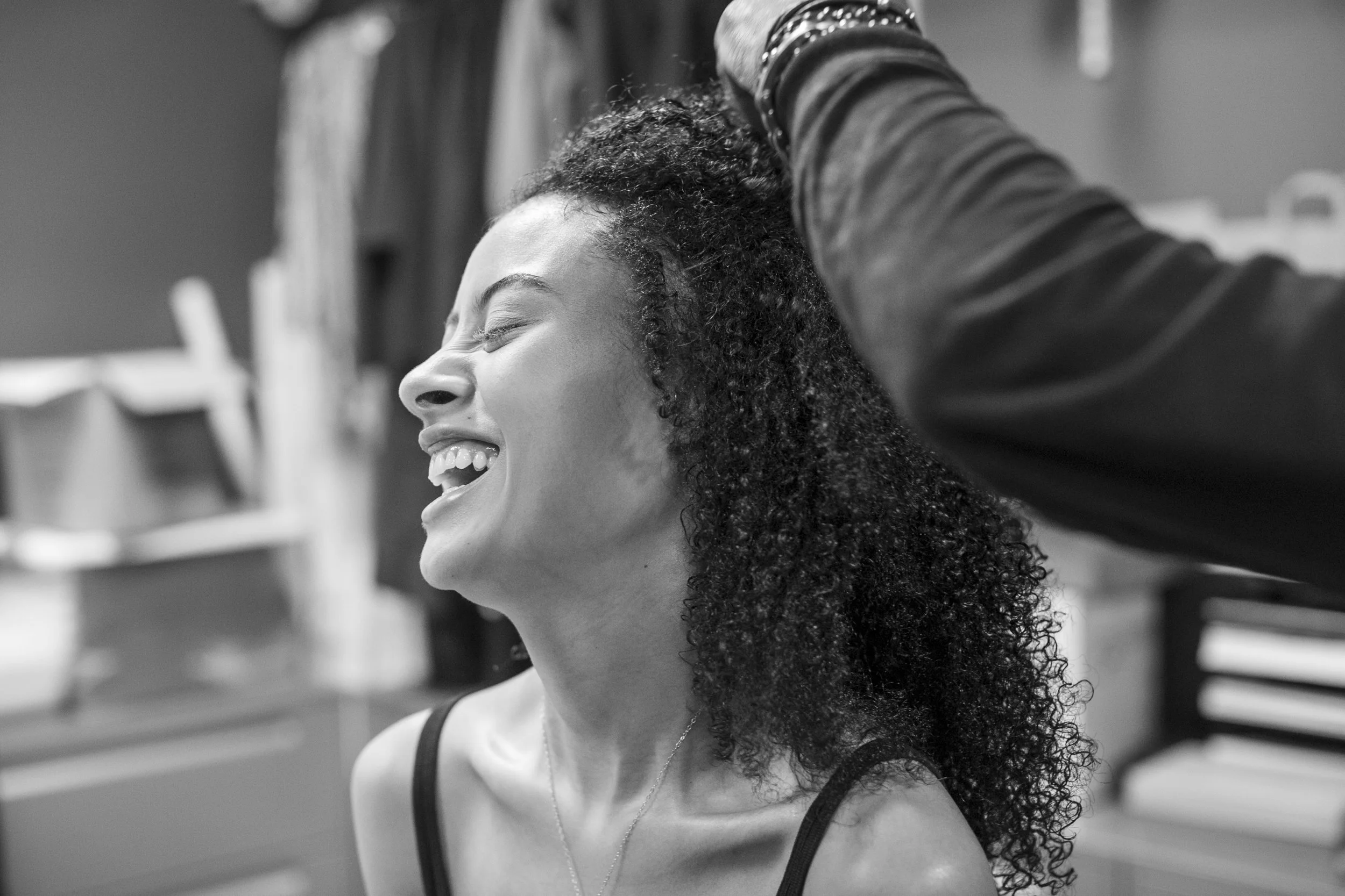 A woman is smiling and laughing while having her hair styled or done by a person whose arm and hand are visible in the image.