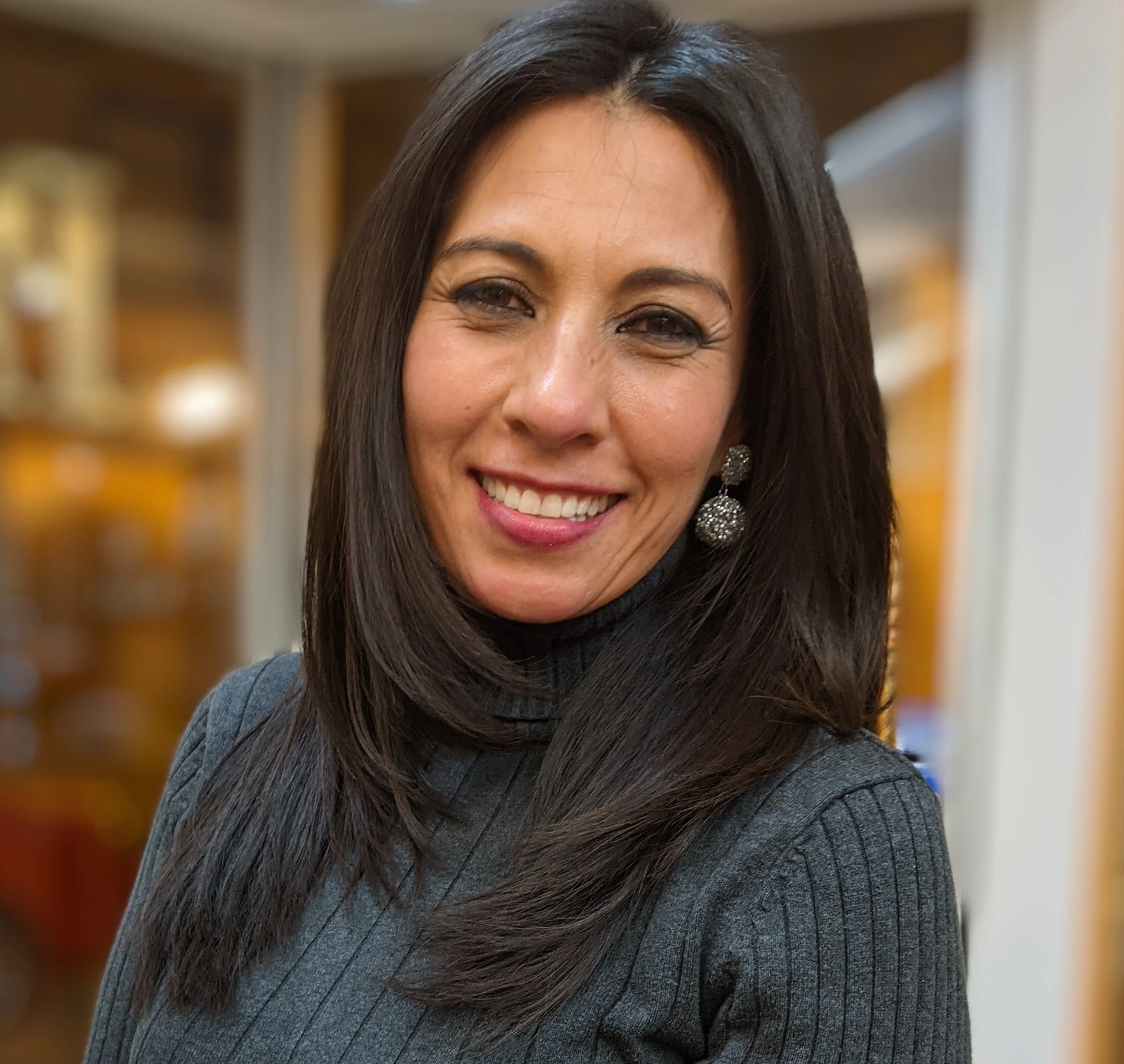 A smiling woman with long dark hair wearing a gray turtleneck sweater and large sparkly earrings, indoors with a blurred background.