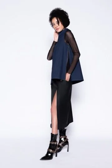 A woman with curly black hair wearing a sheer black long-sleeve top, a blue sleeveless dress with a high slit, black high-heeled boots, standing against a plain white background.