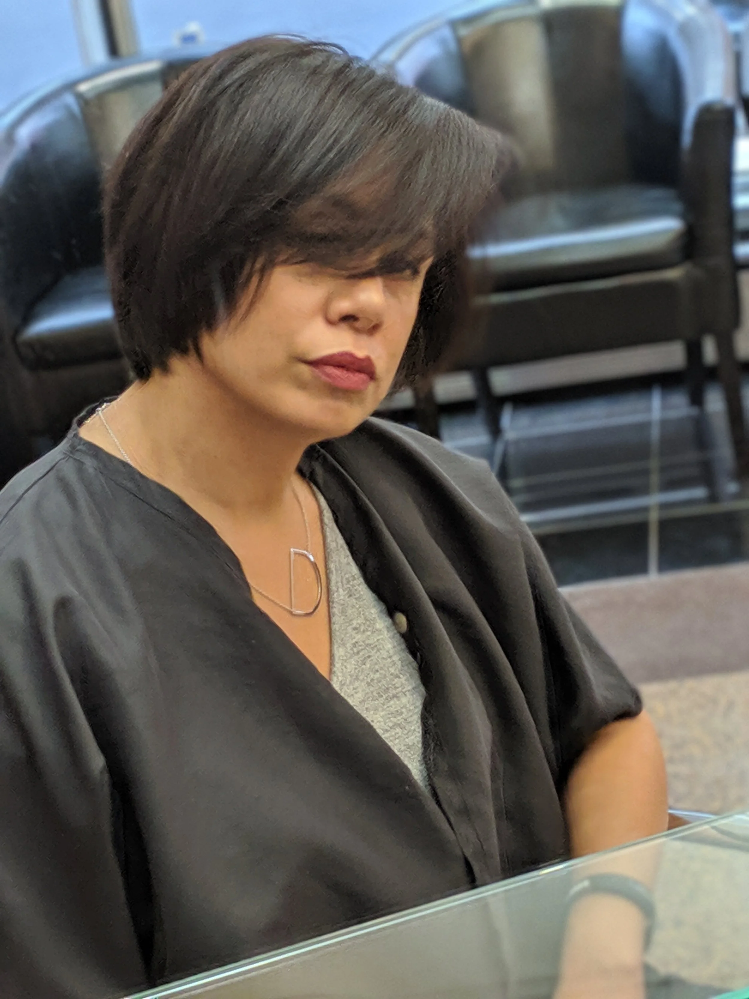 A woman with dark, shoulder-length hair, wearing a gray top and a black jacket, sitting at a table inside a room with black chairs in the background.
