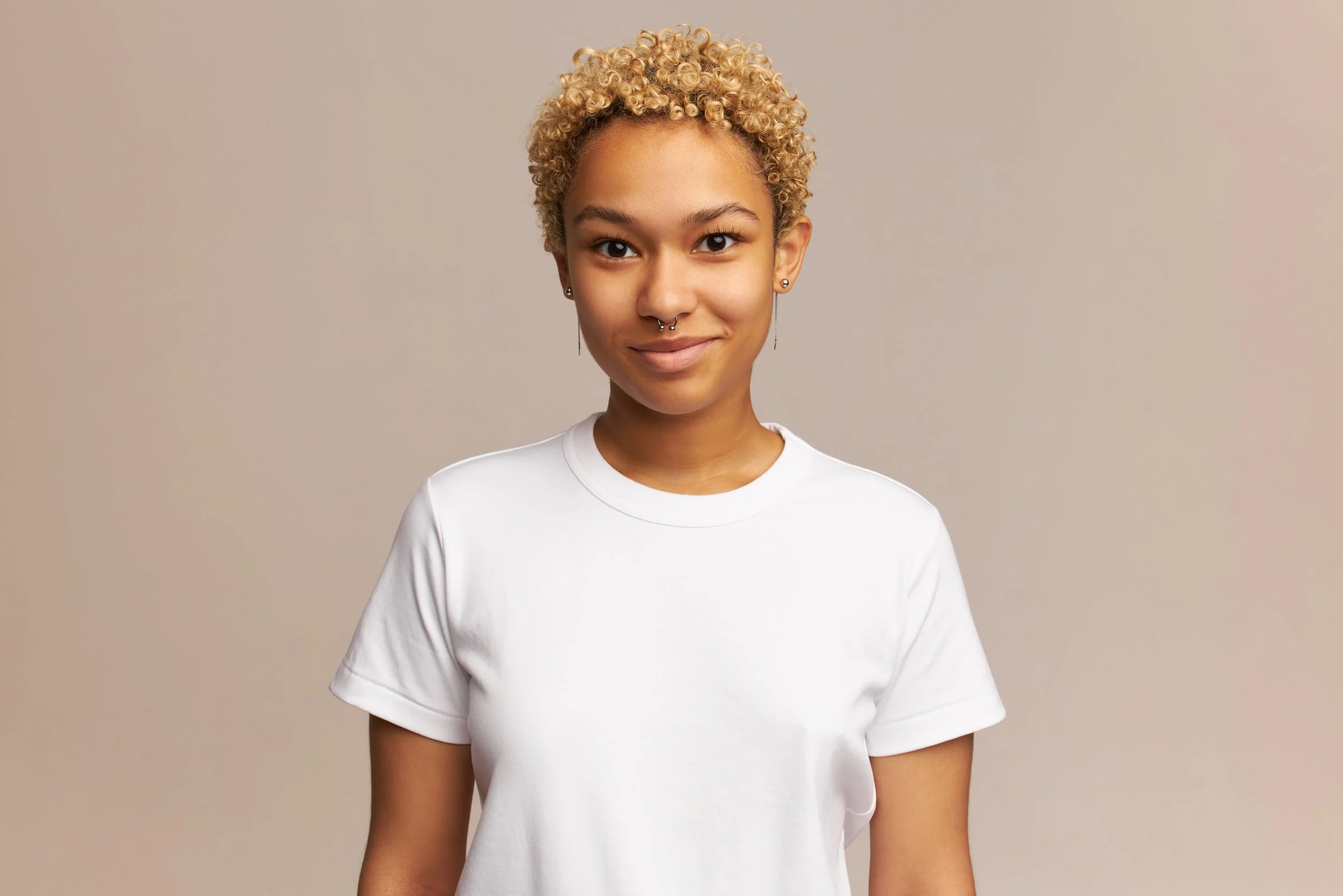 A young woman with short, curly blonde hair, wearing a white t-shirt, standing in front of a plain beige background, smiling slightly at the camera.