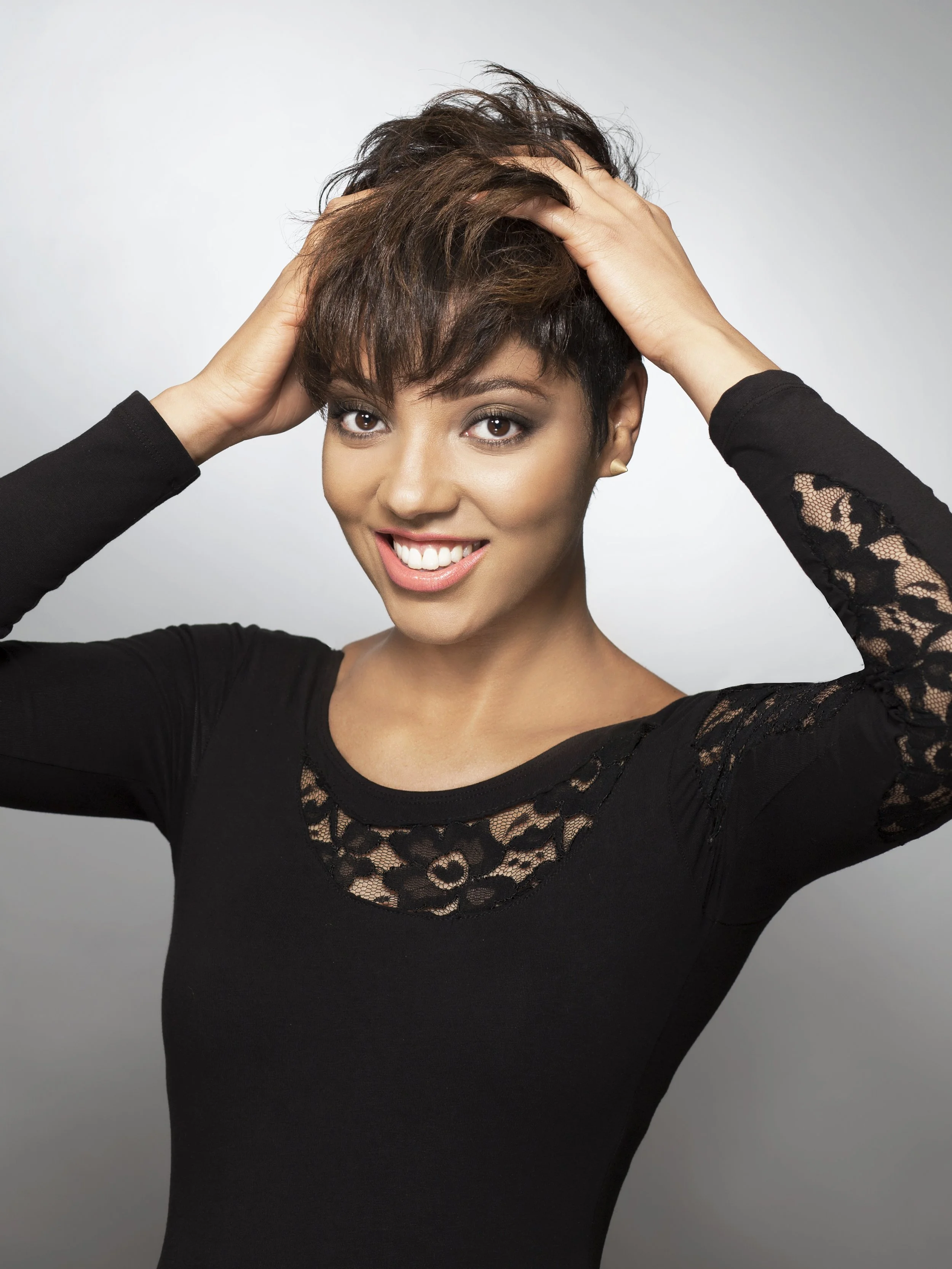 A woman with short, textured brown hair, smiling and touching her hair with both hands. She is wearing a black top with lace details on the sleeves and neckline, and stands against a plain gray background.