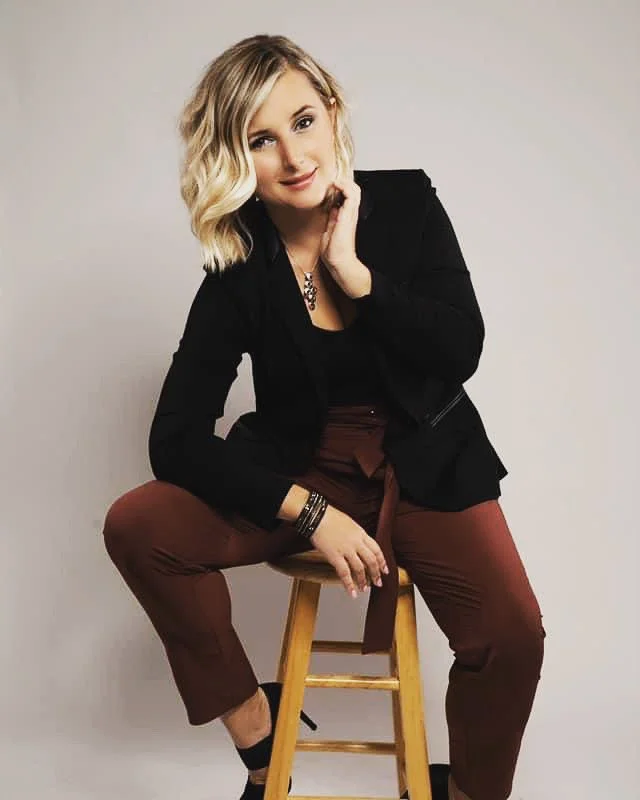 A woman with shoulder-length blonde hair and a black blazer sitting on a wooden stool against a plain white wall.