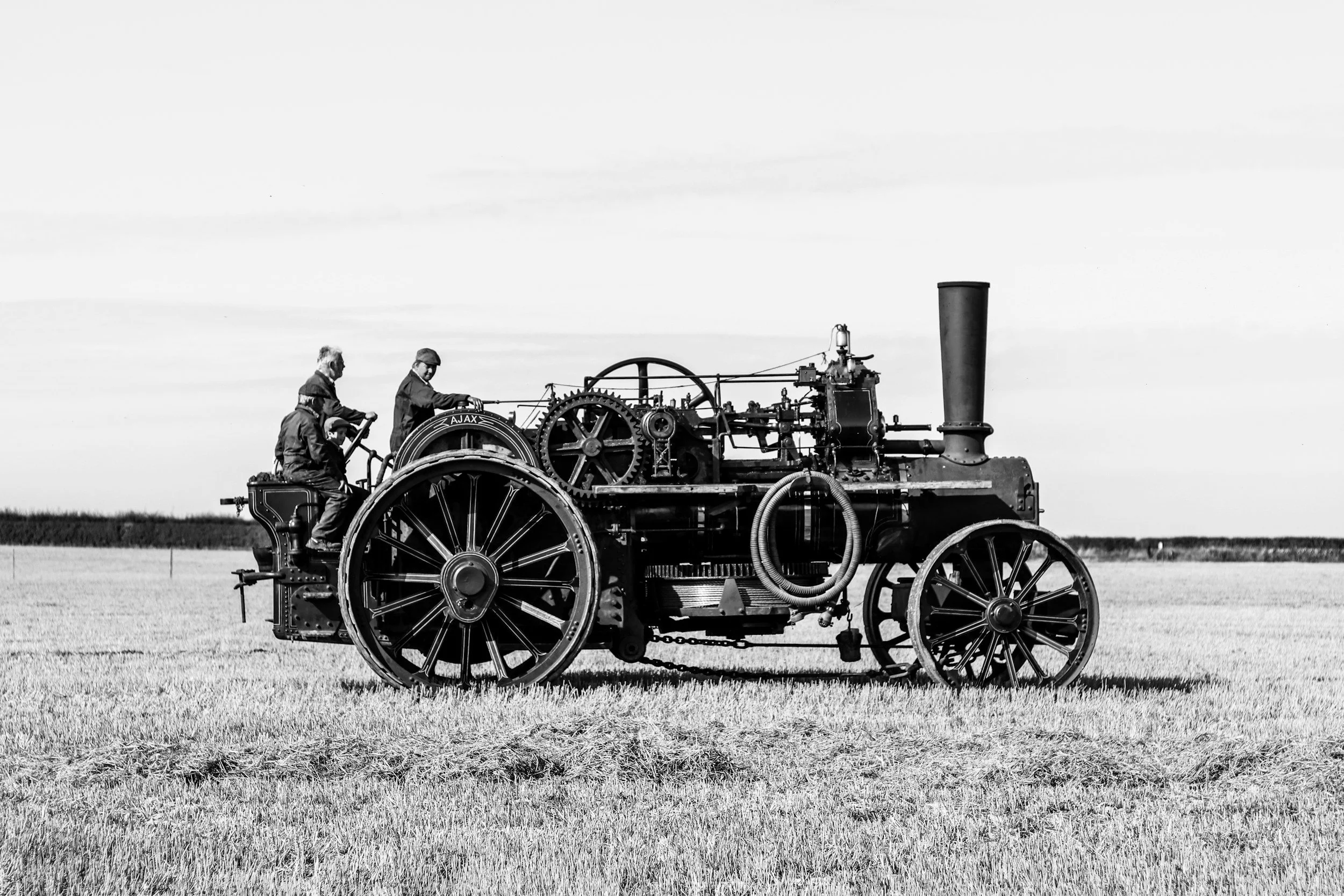 A black and white photo of three people riding an old-fashioned steam-powered tractor in a field, with a flat horizon and cloudy sky in the background.
