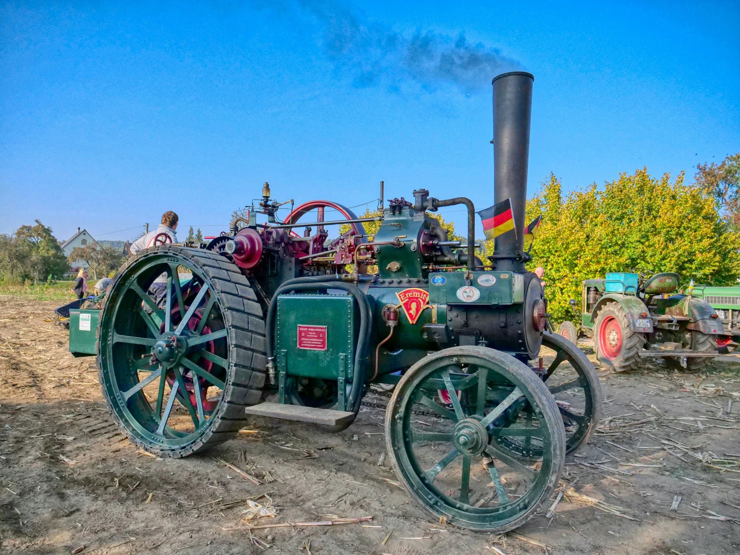 A vintage steam tractor on a dirt field with a second tractor in the background, trees with autumn foliage, and a clear blue sky.
