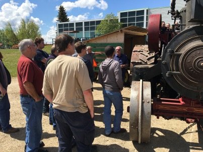 A group of people gathered around a large vintage tractor, listening to a man speaking. The tractor has big wheels and is partly in a rustic setting.