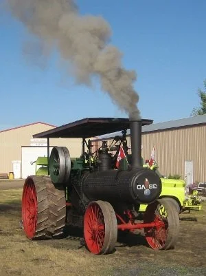 Vintage steam tractor with black boiler, red wheels, and gray smoke coming from chimney, parked on grass near a building.