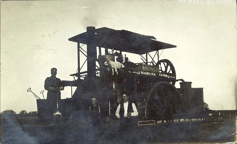 Black and white photograph of a vintage steam tractor on a farm with a dog on top, three men standing nearby, and the farm name 'Namaka Farm' visible on the machinery.