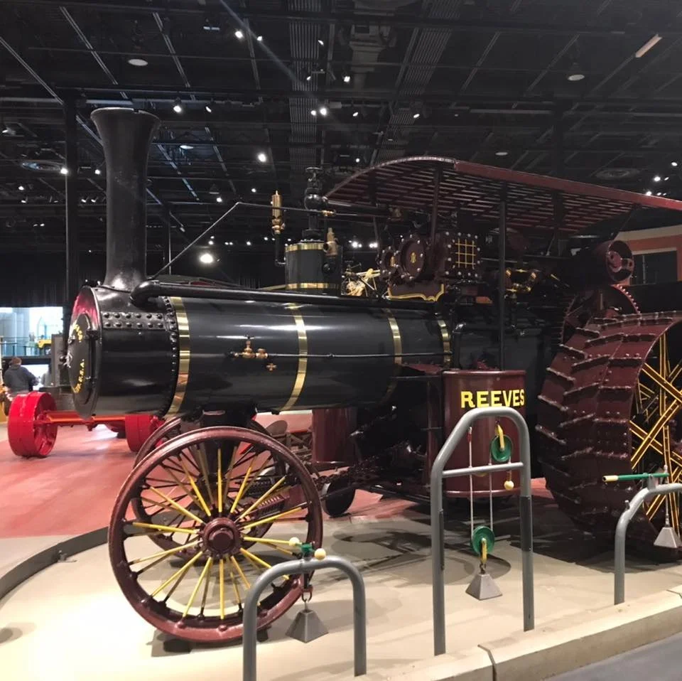 A vintage steam-powered tractor with large tracks on the right and wheels on the left, displayed in a museum with a black ceiling and spotlights.