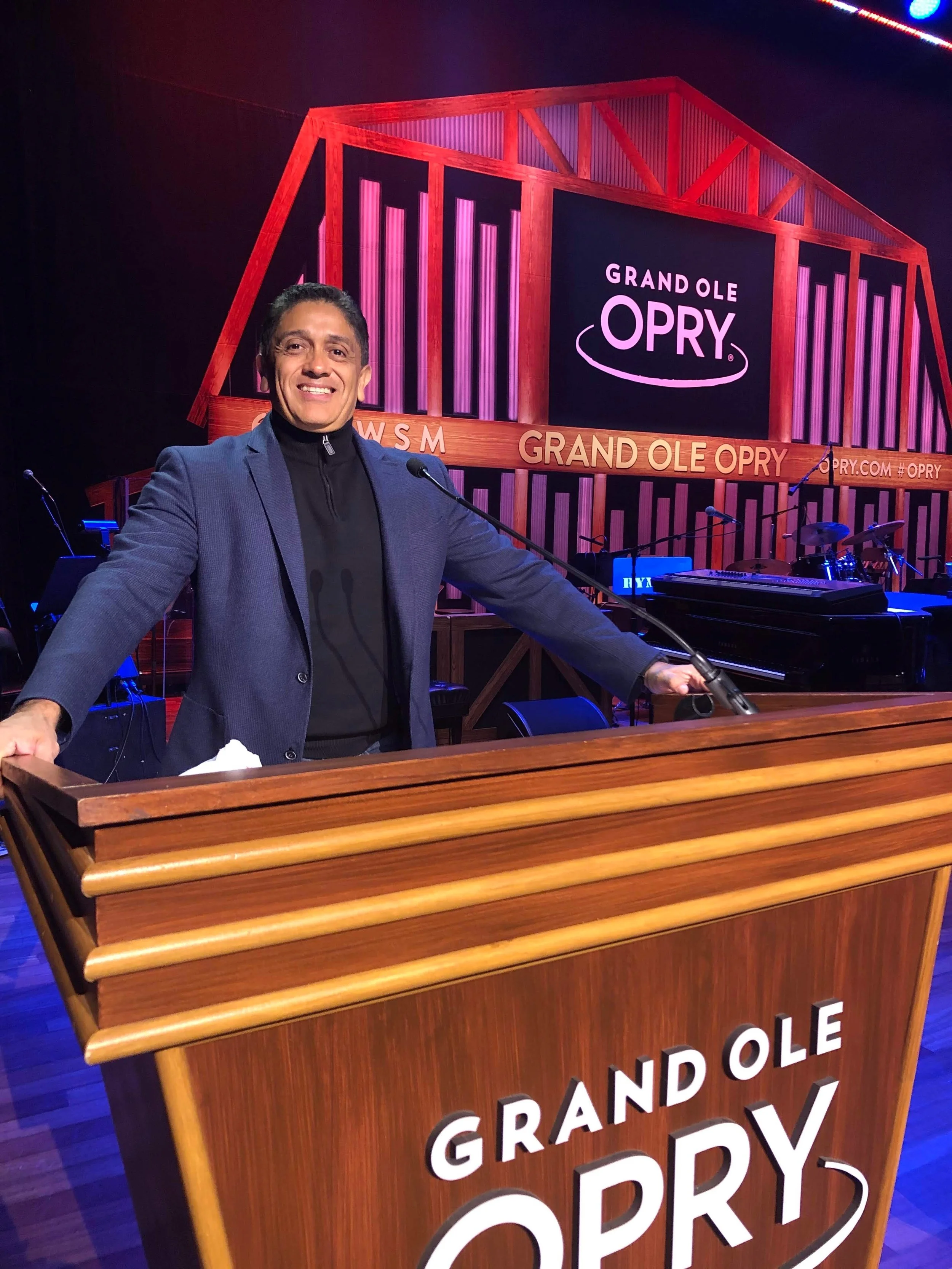 A man standing behind a wooden podium with the words 'Grand Ole Opry' on it, smiling and holding onto the podium at the Grand Ole Opry stage, with the stage backdrop displaying the Opry logo and the name 'Grand Ole Opry'.