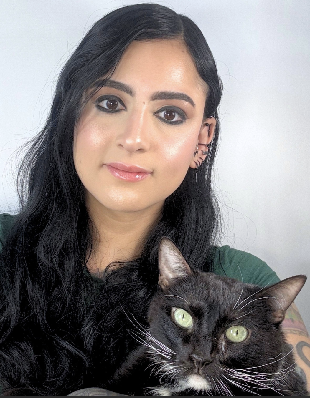 A woman with dark hair and makeup holding a black cat with green eyes.