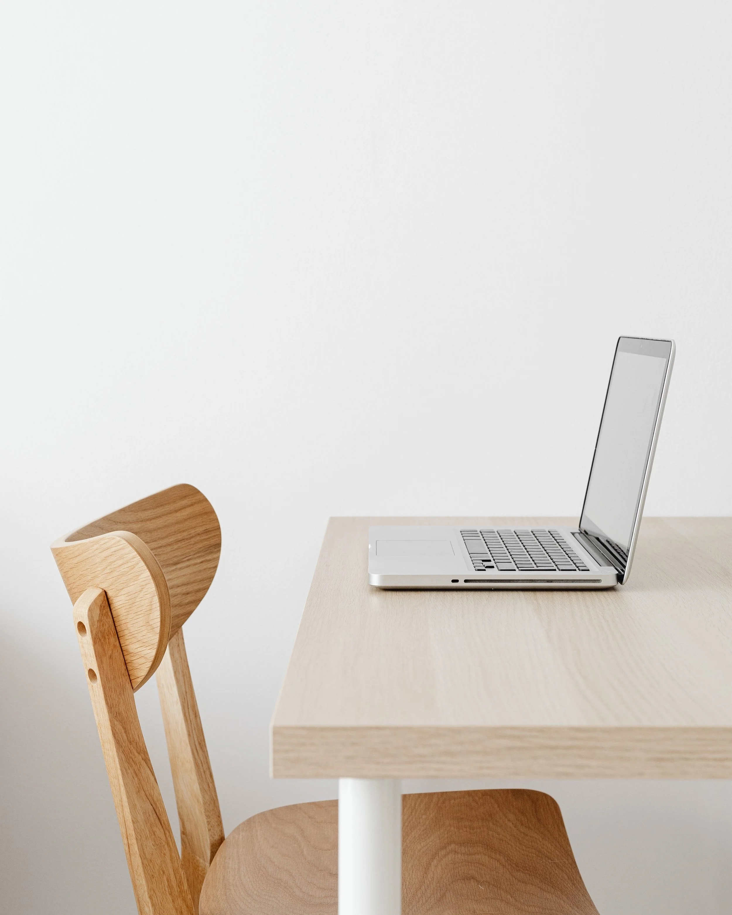 A minimalist workspace with a light wood desk, a wooden chair, and an open silver laptop on the desk against a plain white wall.