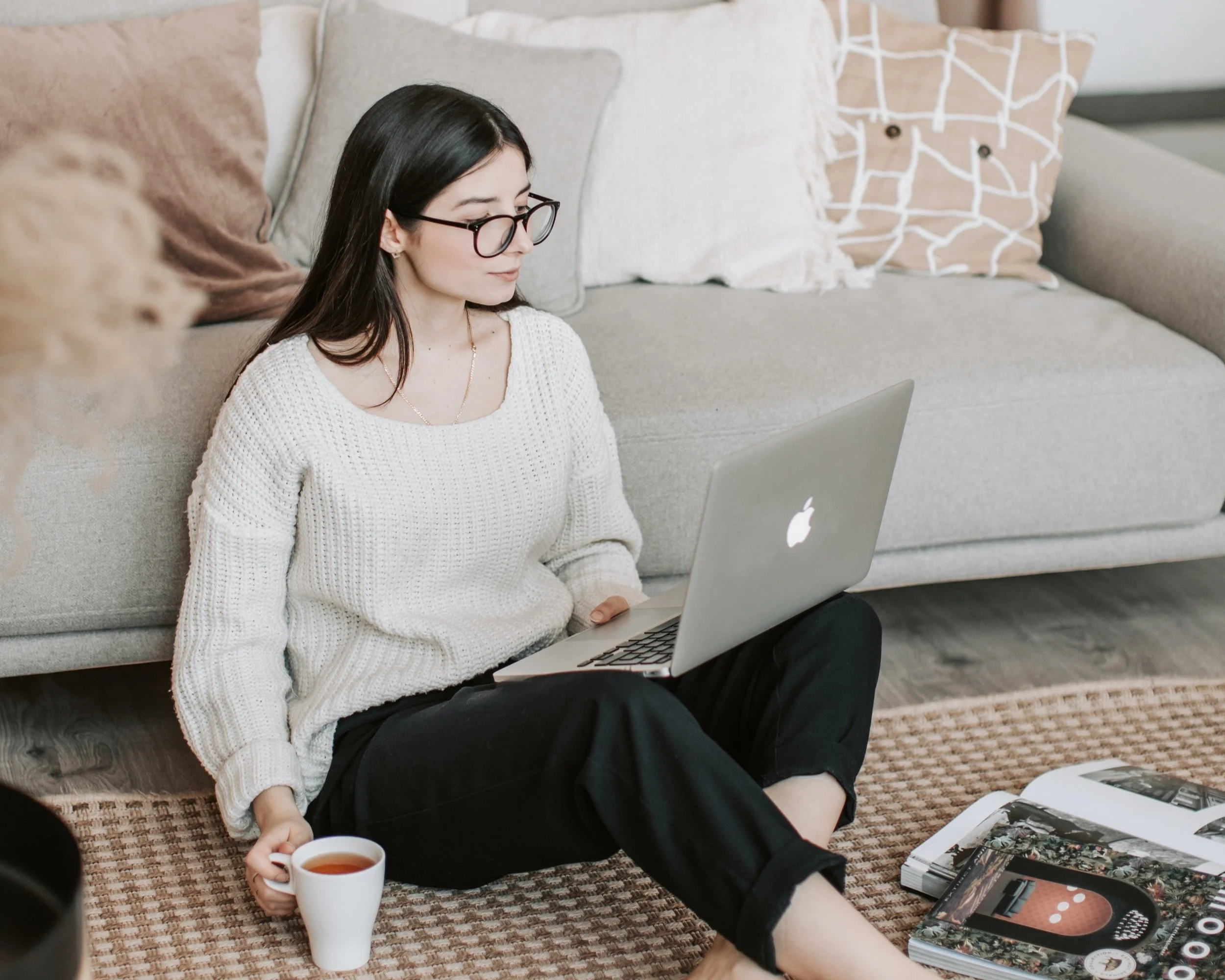 A woman with long dark hair, glasses, and a white sweater sitting on the floor with a laptop on her lap, a cup of tea in her hand, and magazines spread out on the rug in front of her, in a cozy living room.