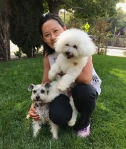 A woman smiling outdoors holding a fluffy white poodle and standing next to a small dog on the grass.
