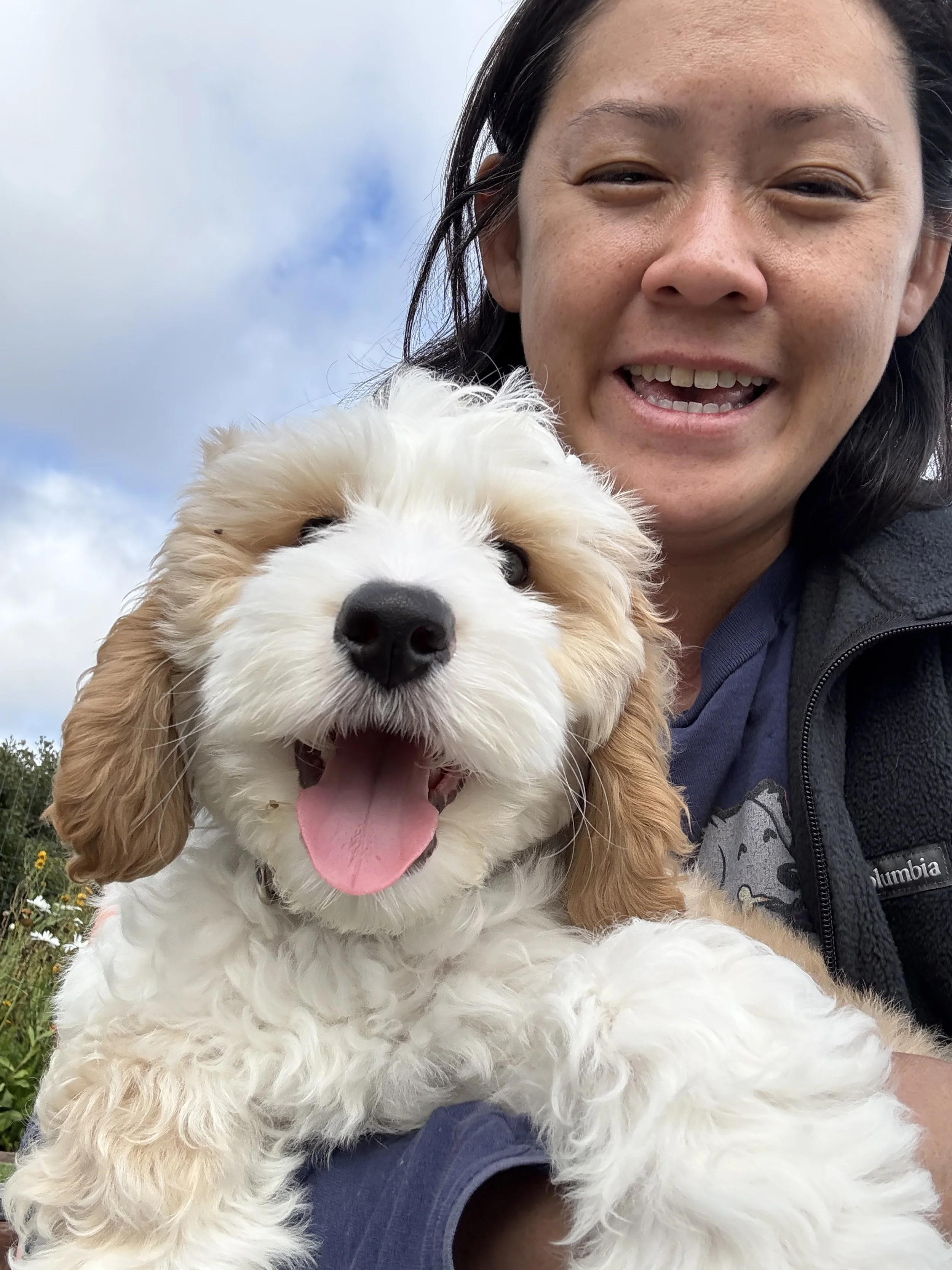 A woman smiling with a happy, fluffy, white and tan puppy, outdoors with a partly cloudy sky in the background.