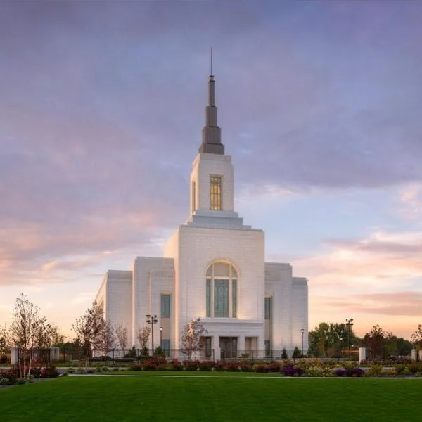 A large white temple building with a tall spire, surrounded by a landscaped area with grass and flowers, set against a sunset sky.