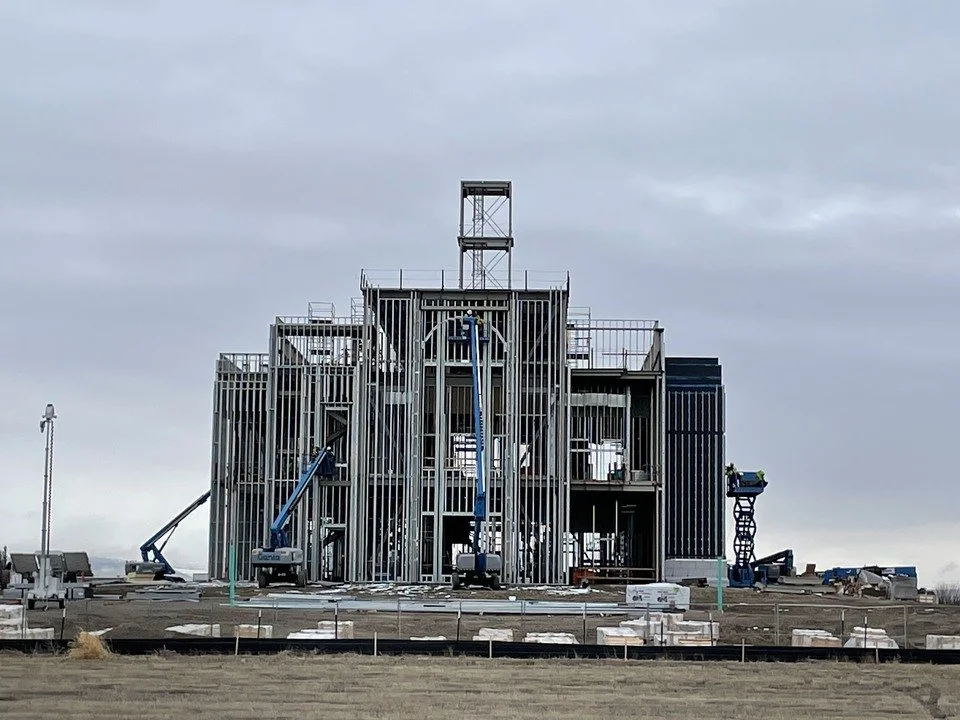 Construction site showing the Burley Idaho Temple building framework made of metal beams with workers using lifts, under a cloudy sky.