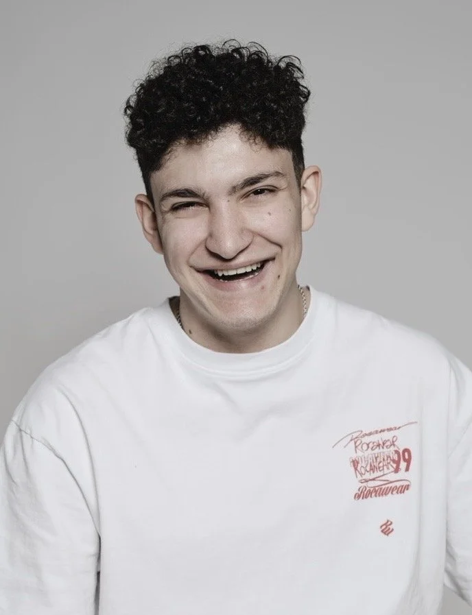 A young man with curly dark hair smiling, wearing a white T-shirt with red text and graphics, against a gray background.