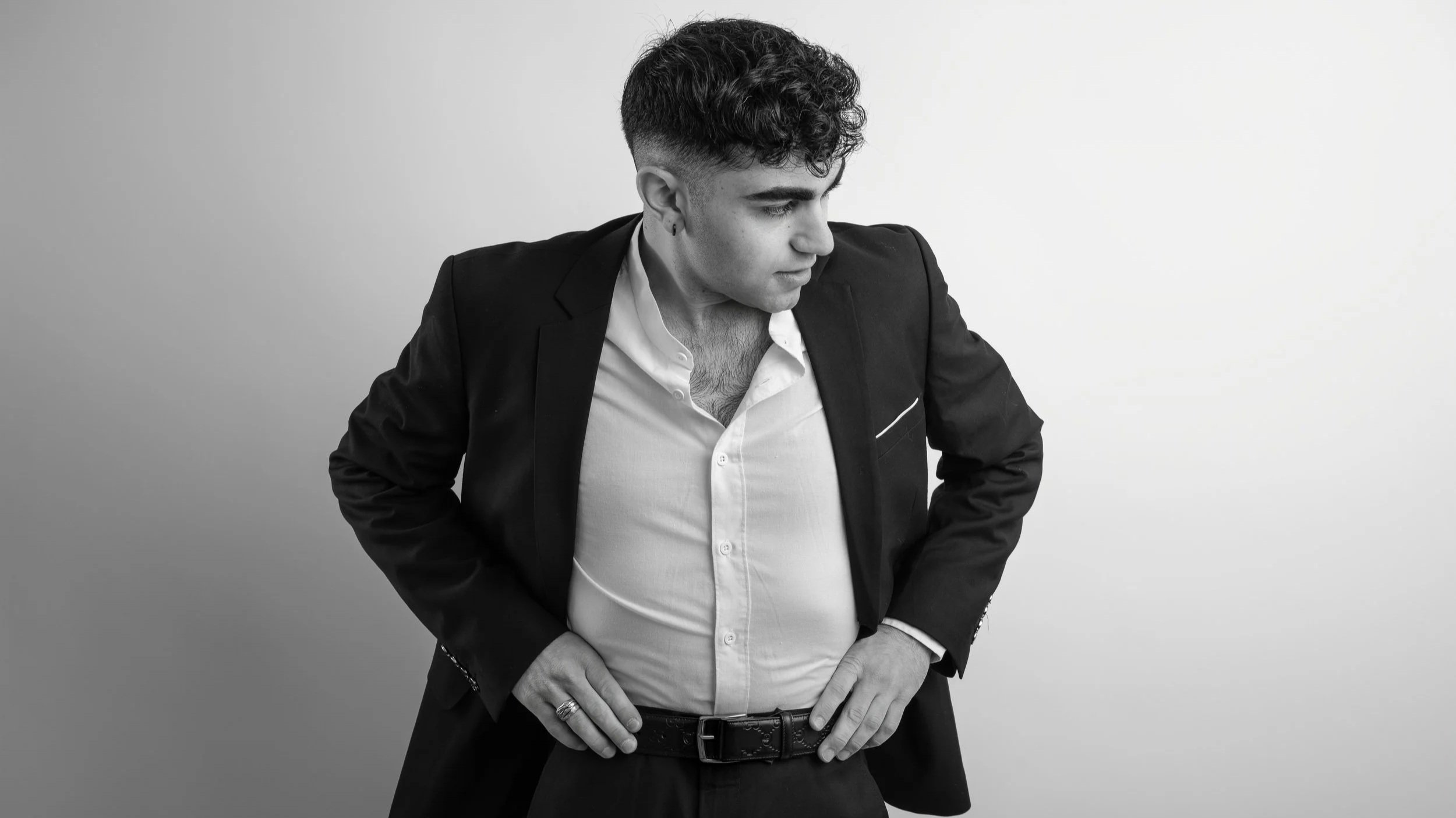 Black and white portrait of a young man with dark curly hair, wearing an open collared shirt and a blazer, standing against a plain background with his hands on his hips, looking to his right.