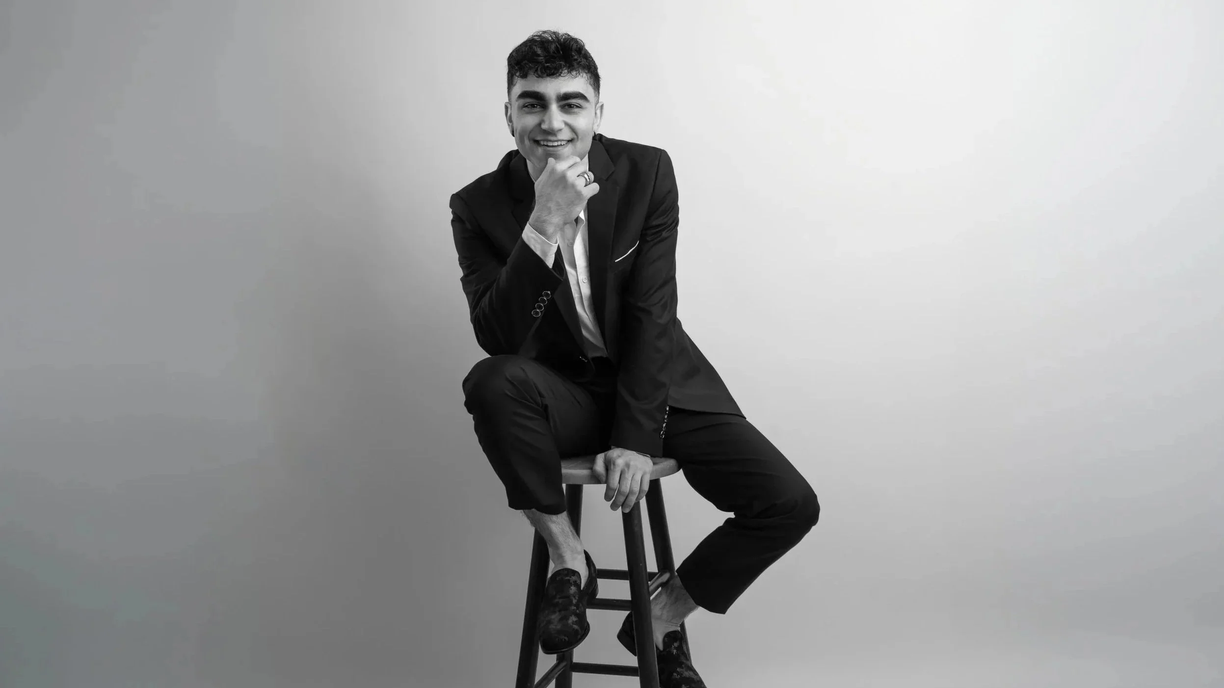 A young man sitting on a stool, wearing a suit and smiling with his chin resting on his hand, in a black-and-white photo against a plain background.