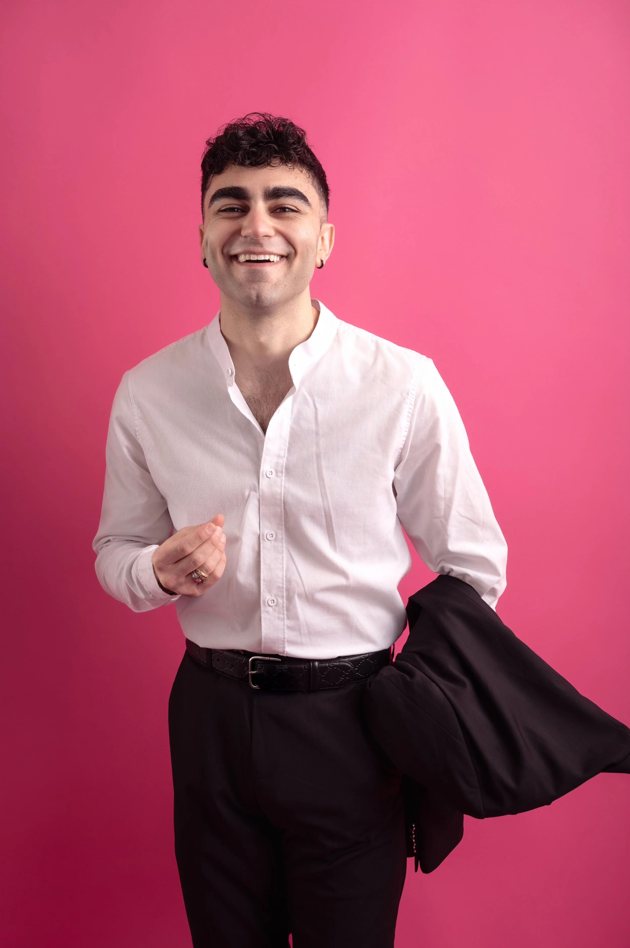 A smiling young man with dark curly hair, wearing a white dress shirt, is standing against a solid pink background, holding a black jacket in his right hand.
