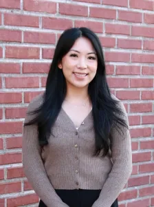 A young woman with long black hair standing in front of a red brick wall, smiling and wearing a beige cardigan.
