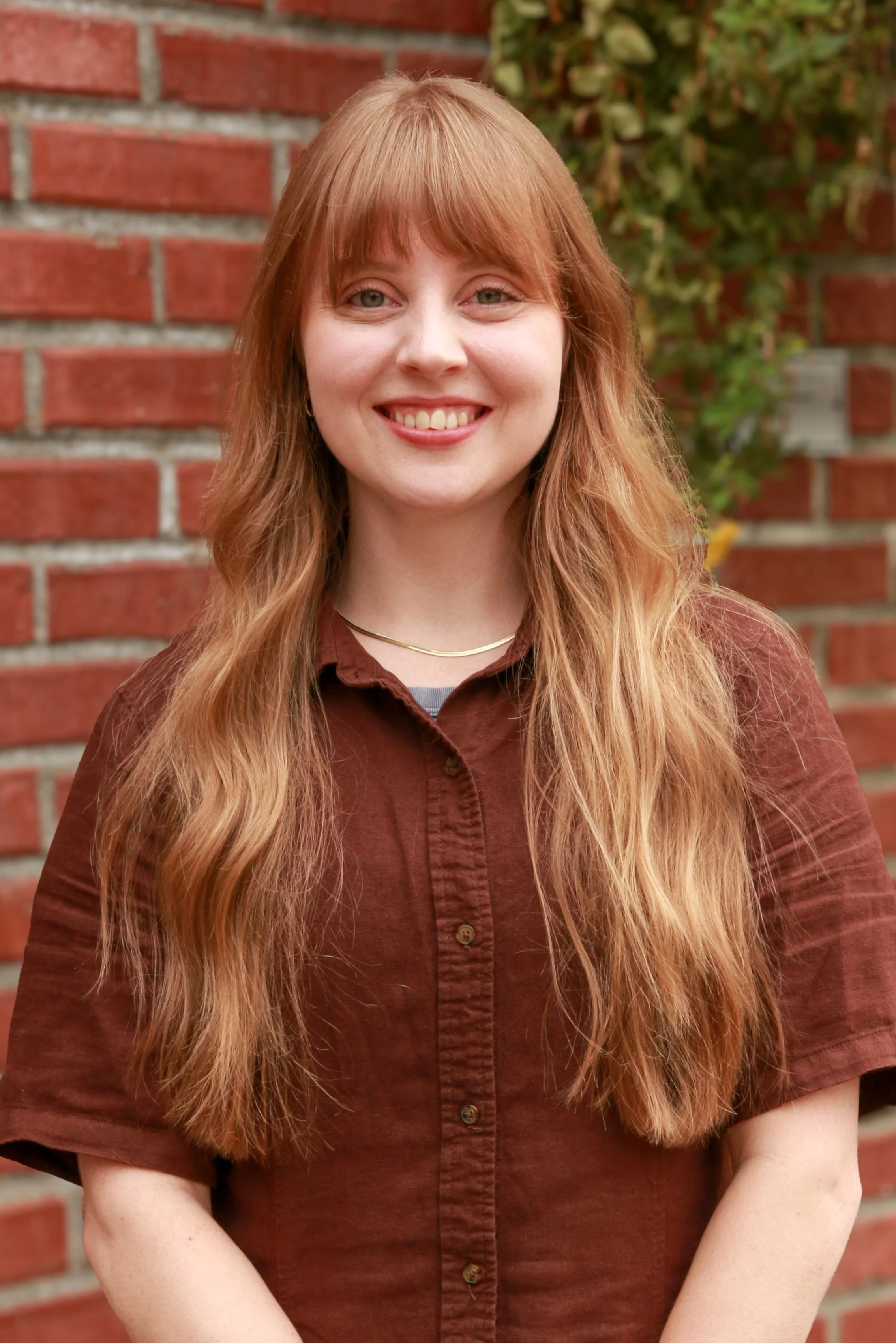 Portrait of a young woman with long red hair smiling, wearing a brown button-up shirt, standing in front of a brick wall with greenery.