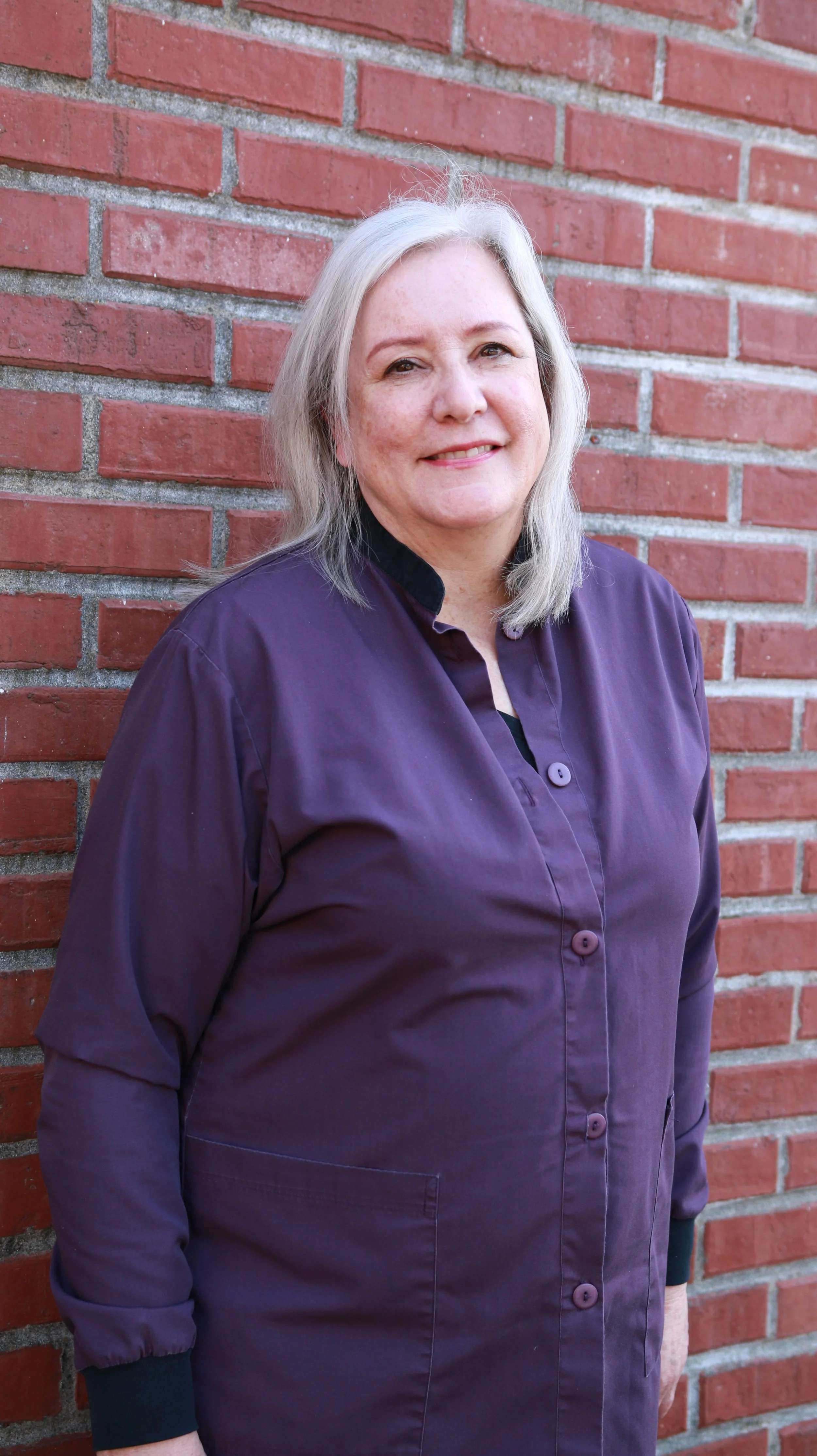 A woman with blonde hair standing in front of a brick wall, wearing a purple shirt and smiling.