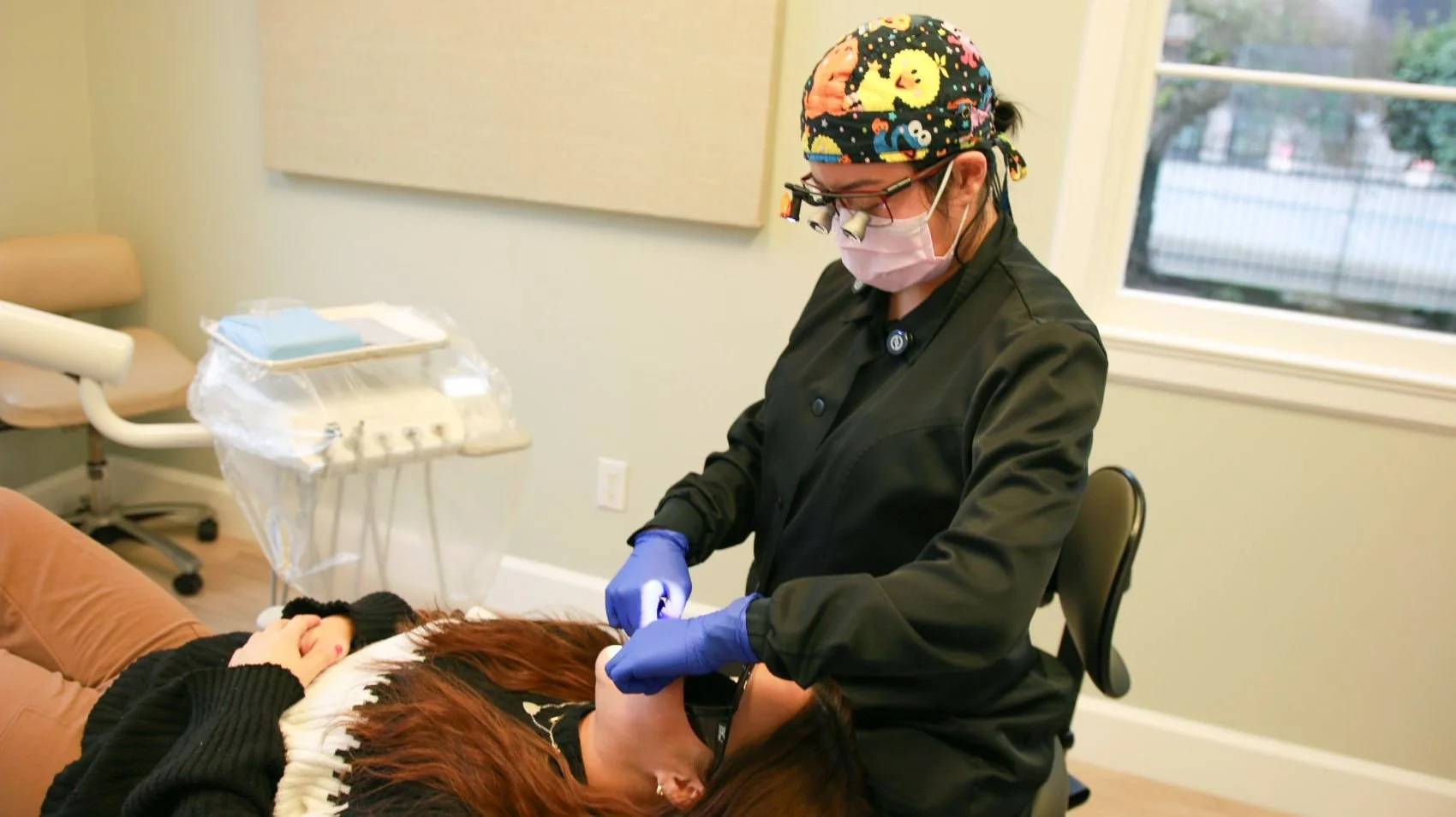 Dentist wearing colorful scrub cap, face mask, and gloves treating a patient lying on dental chair.